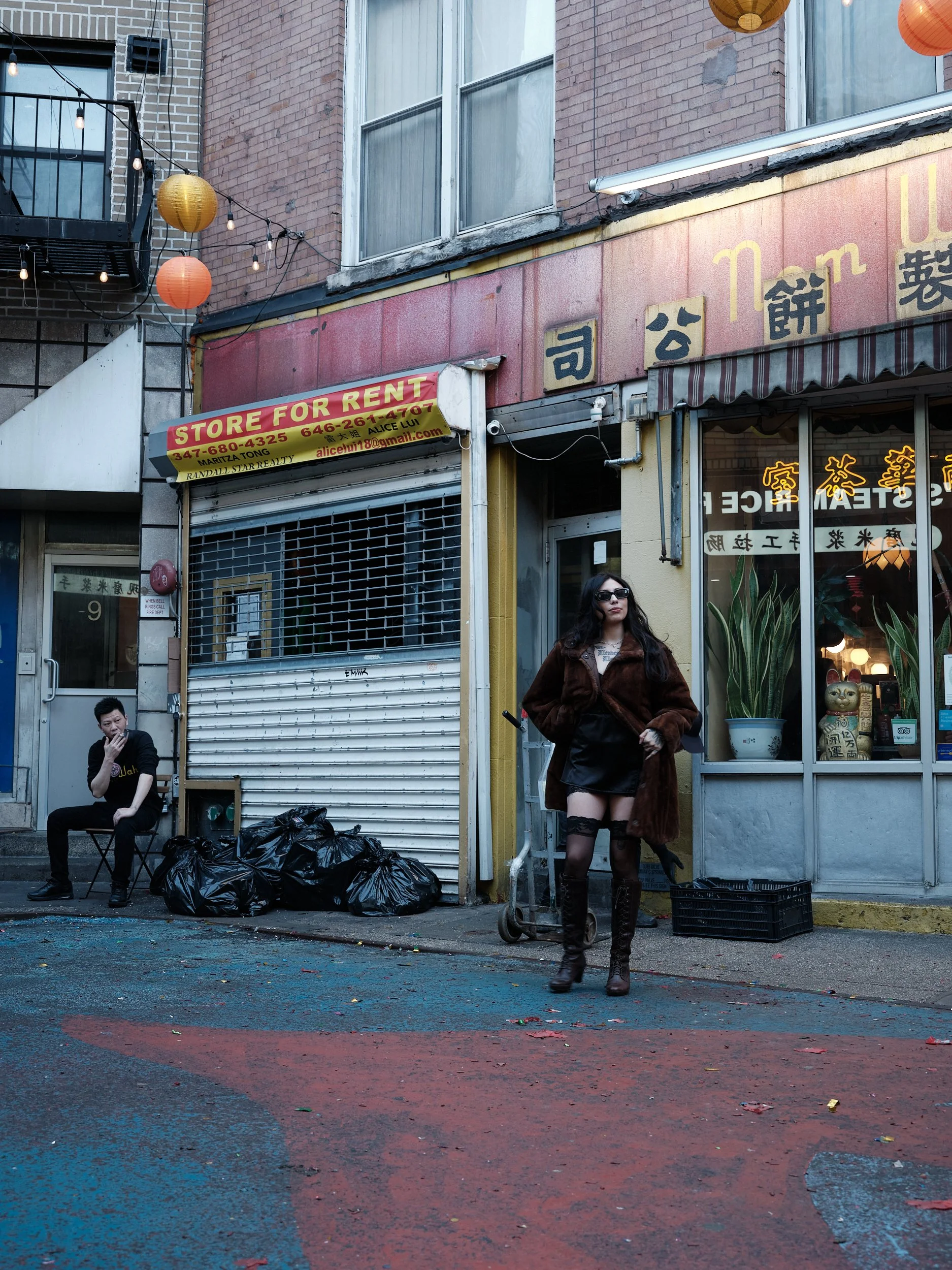 A woman in sunglasses and thigh-high boots standing outside a storefront on a city street sidewalk. A man is sitting on a chair nearby with plastic bags on the ground beside him. The storefront has a sign that reads 'STORE FOR RENT' and a window with