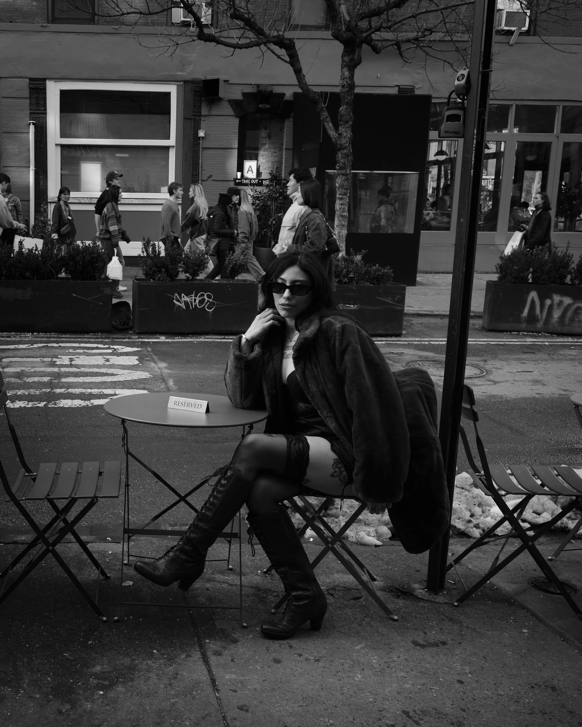 A woman with dark hair, wearing sunglasses, a fuzzy jacket, thigh-high stockings, and high-heeled boots, sitting at an outdoor cafe table marked reserved. The scene is in black and white, with a city street, pedestrians, buildings, and a tree in the 
