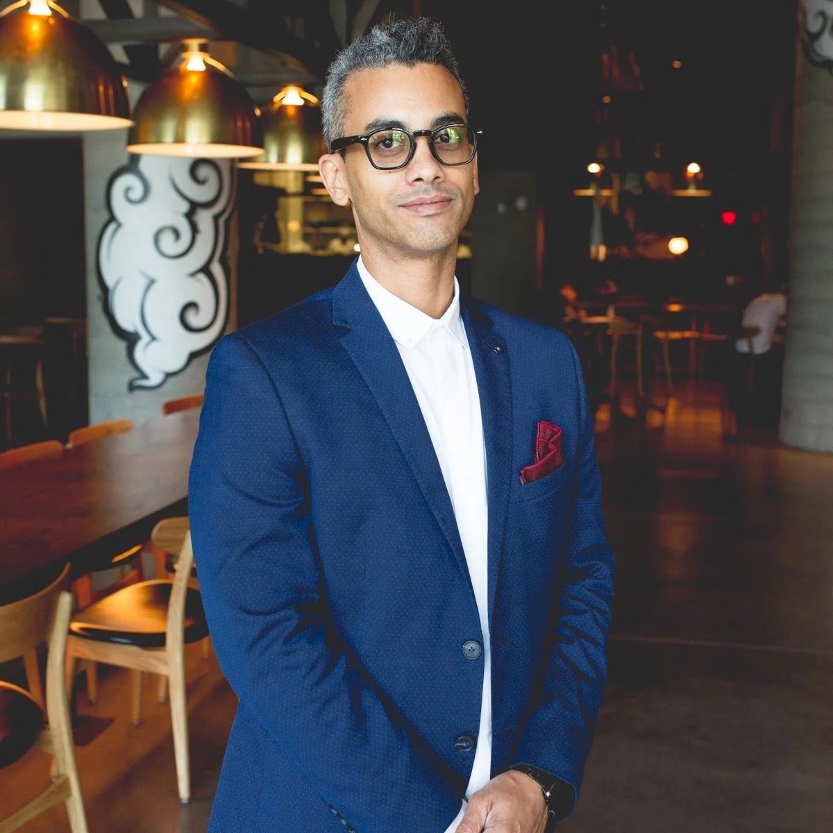 A man in a blue suit and glasses standing in a restaurant with wooden tables and chairs, modern lighting, and artwork on the wall.