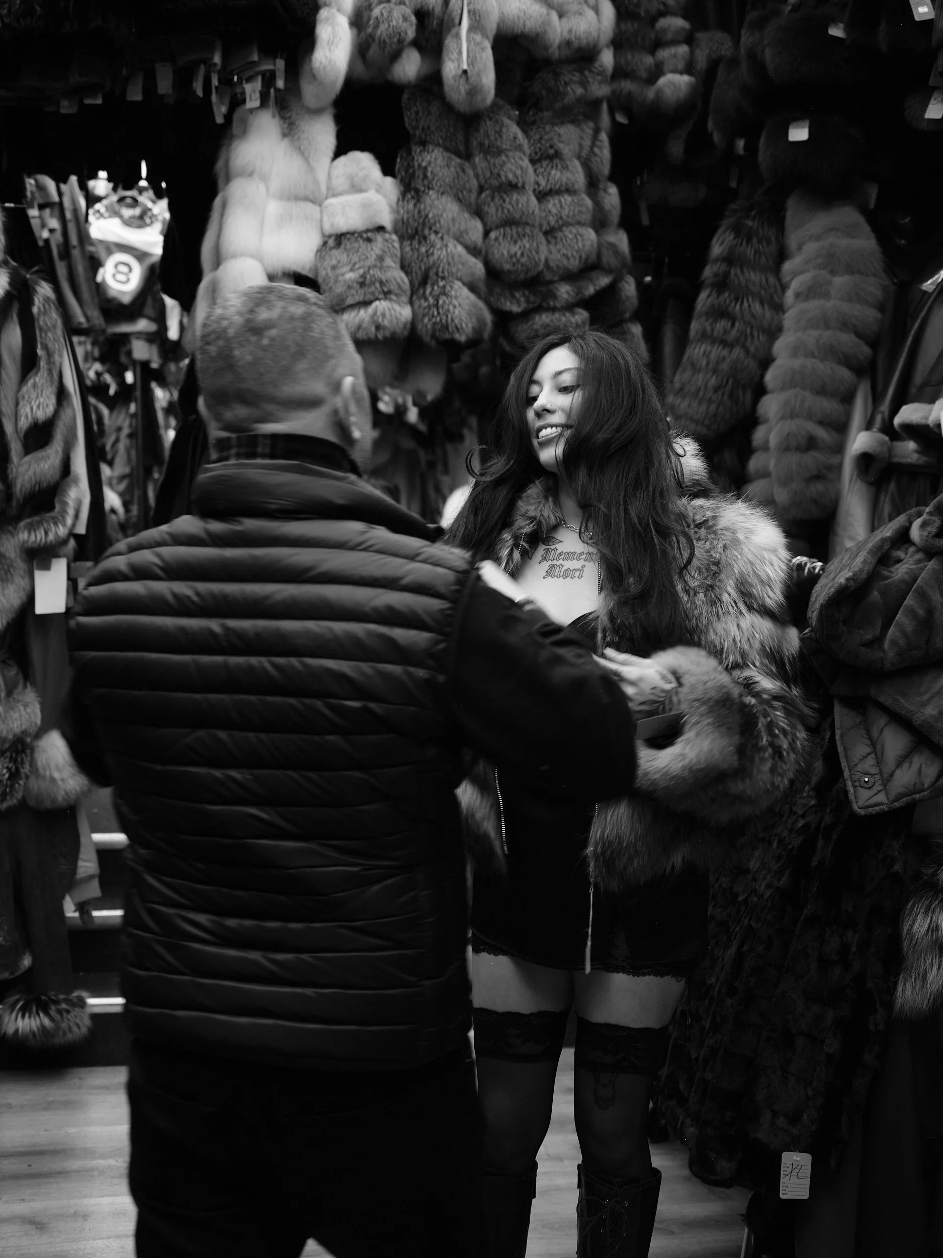 A woman with long dark hair, wearing a fur coat, smiling, and a man in a puffer jacket, shopping for fur coats in a store.