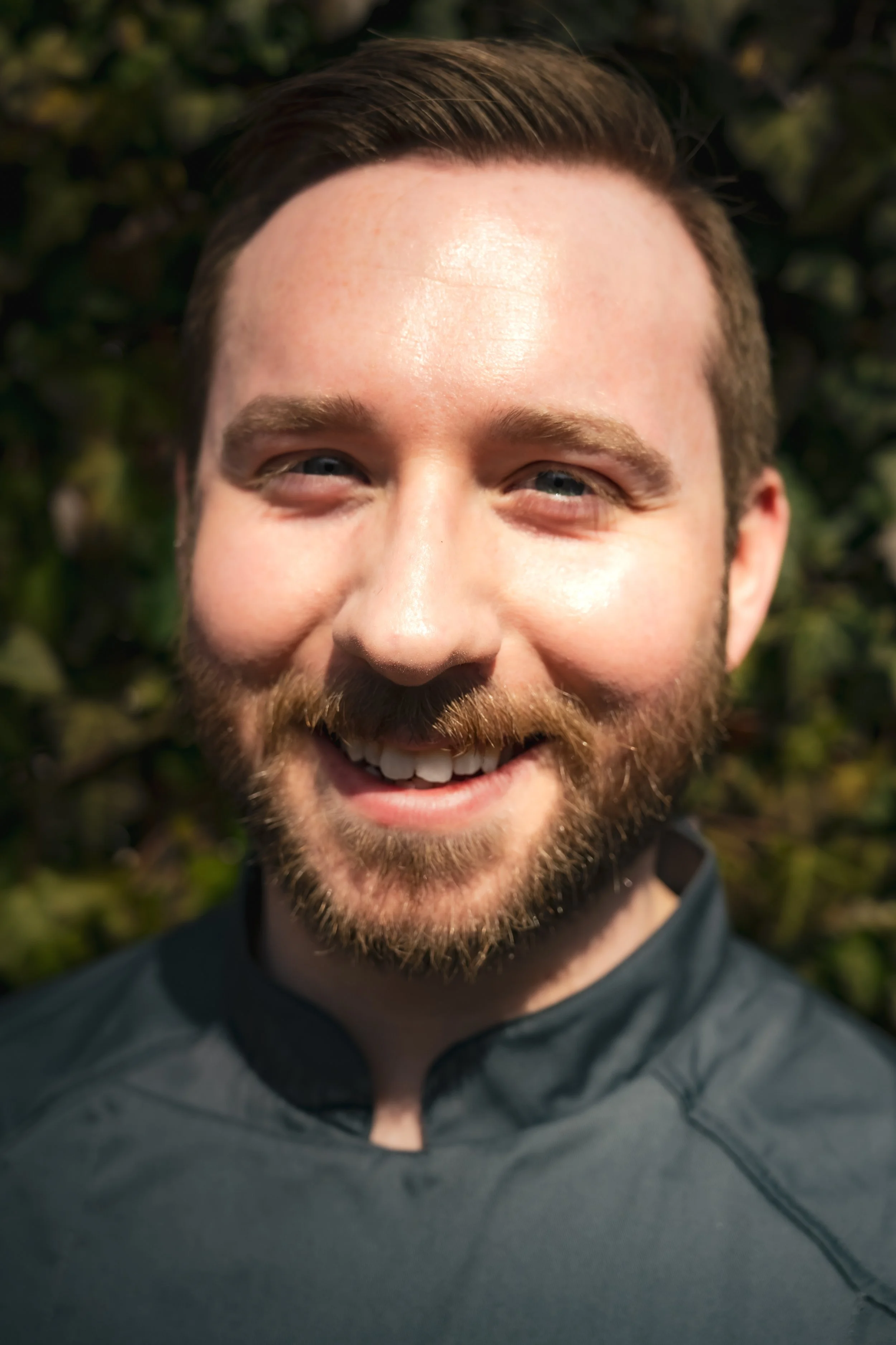 Chef portrait, wearing a black athletic shirt, outdoors with green foliage in the background.