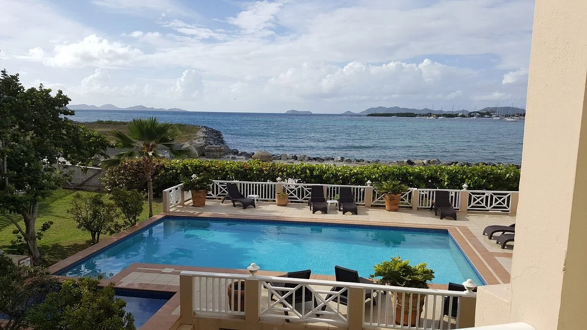 View of a swimming pool overlooking the ocean, with lounge chairs, potted plants, and a white fence in a tropical setting.