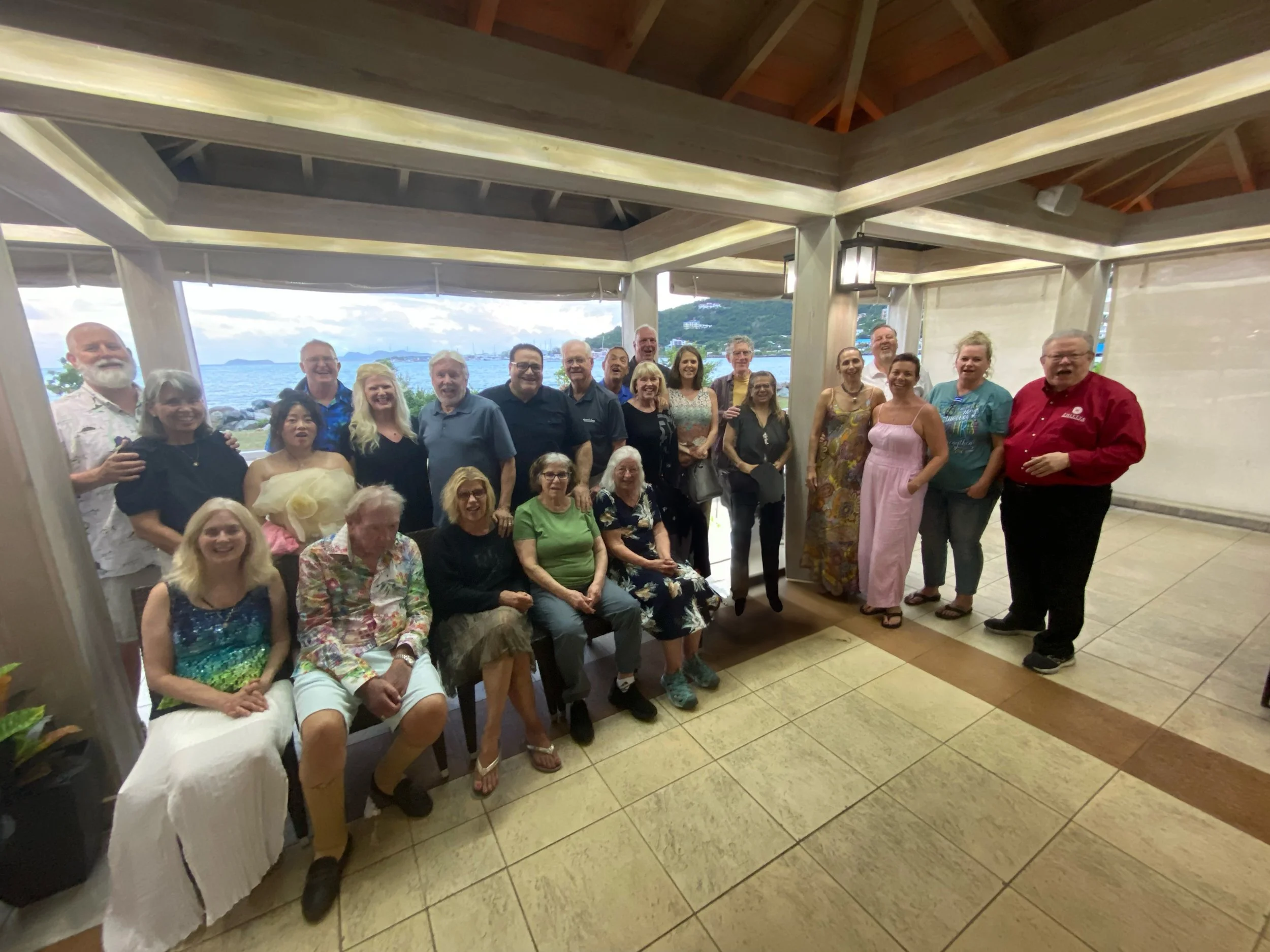 A group of around 25 people posing for a photo inside a covered area with large windows showing a scenic ocean view in the background. The group is smiling and appears happy, dressed in casual and semi-formal clothing. Some are seated, and others are standing.