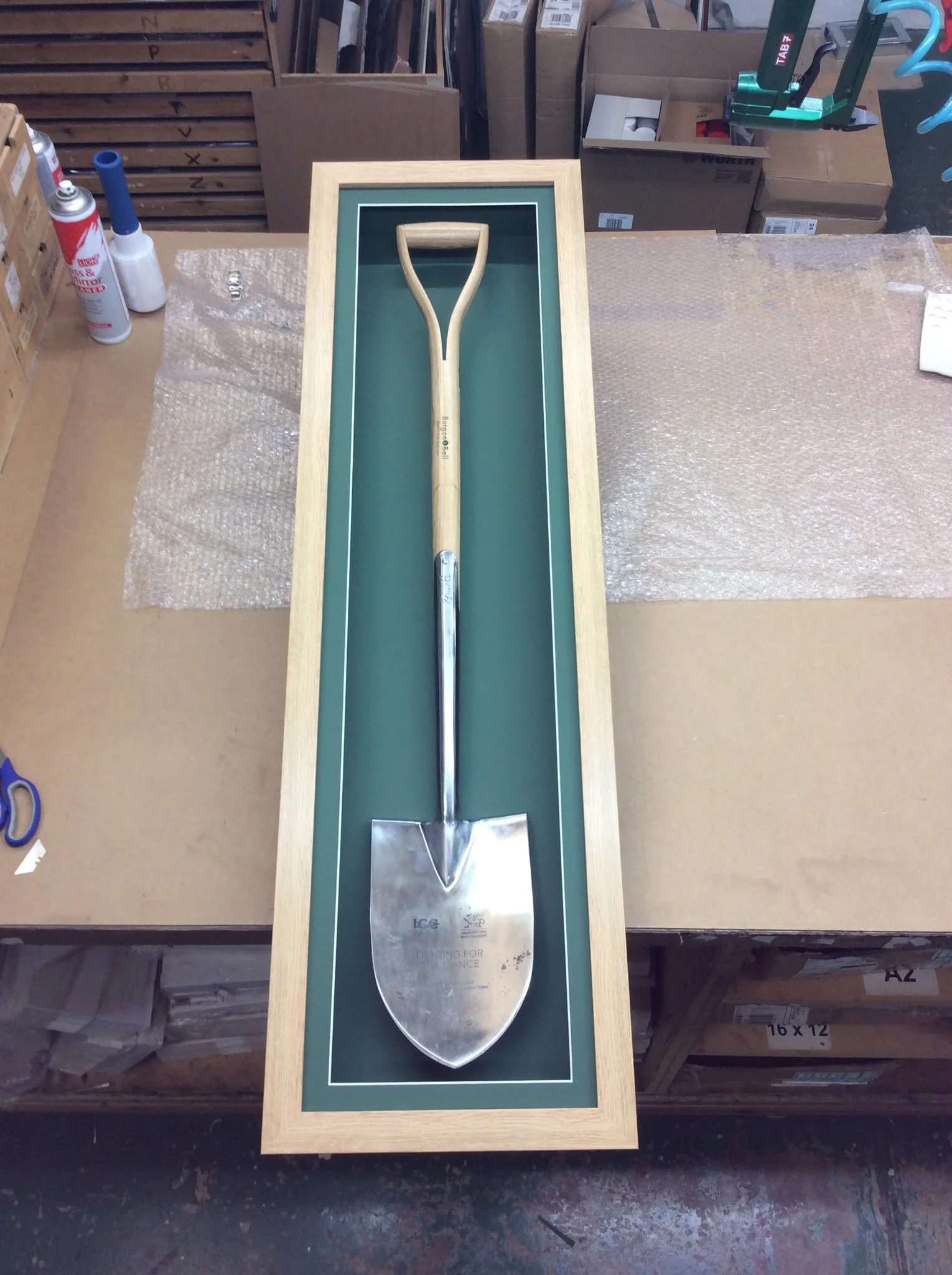 A metal shovel with a wooden handle displayed inside a green and wooden frame on a workspace table.