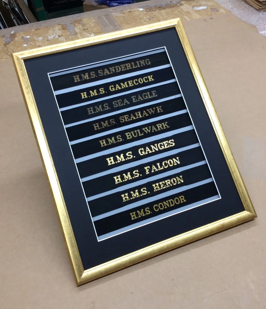 Framed display listing the names of various historic ships, including H.M.S. Sanderling, H.M.S. Gamecock, H.M.S. Sea Eagle, H.M.S. Seahawk, H.M.S. Bulwark, H.M.S. Ganges, H.M.S. Falcon, H.M.S. Heron, and H.M.S. Condor.