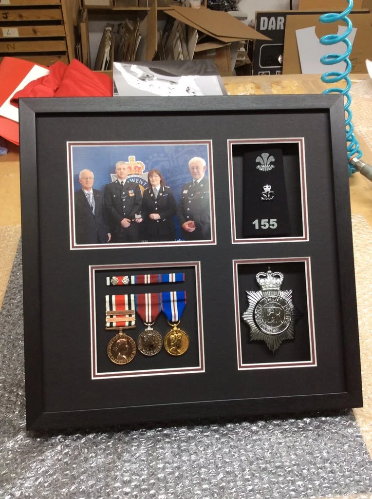 A framed display containing a photograph of four police officers, three medals, two insignias, and a badge, all commemorating police service. The display is on a workbench in a workshop environment.