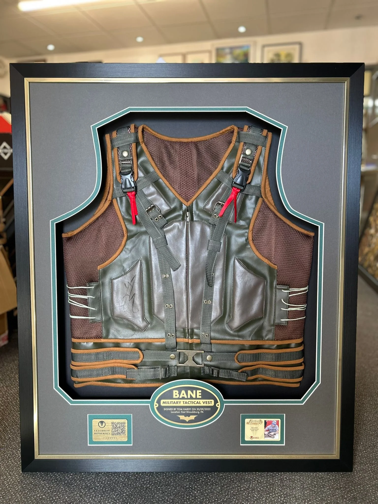 Display case with a framed, signed military tactical vest labeled 'Bane'.