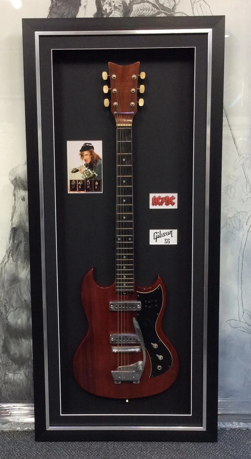 A vintage Gibson SG electric guitar displayed in a black shadow box with AC/DC and band member images and stickers.