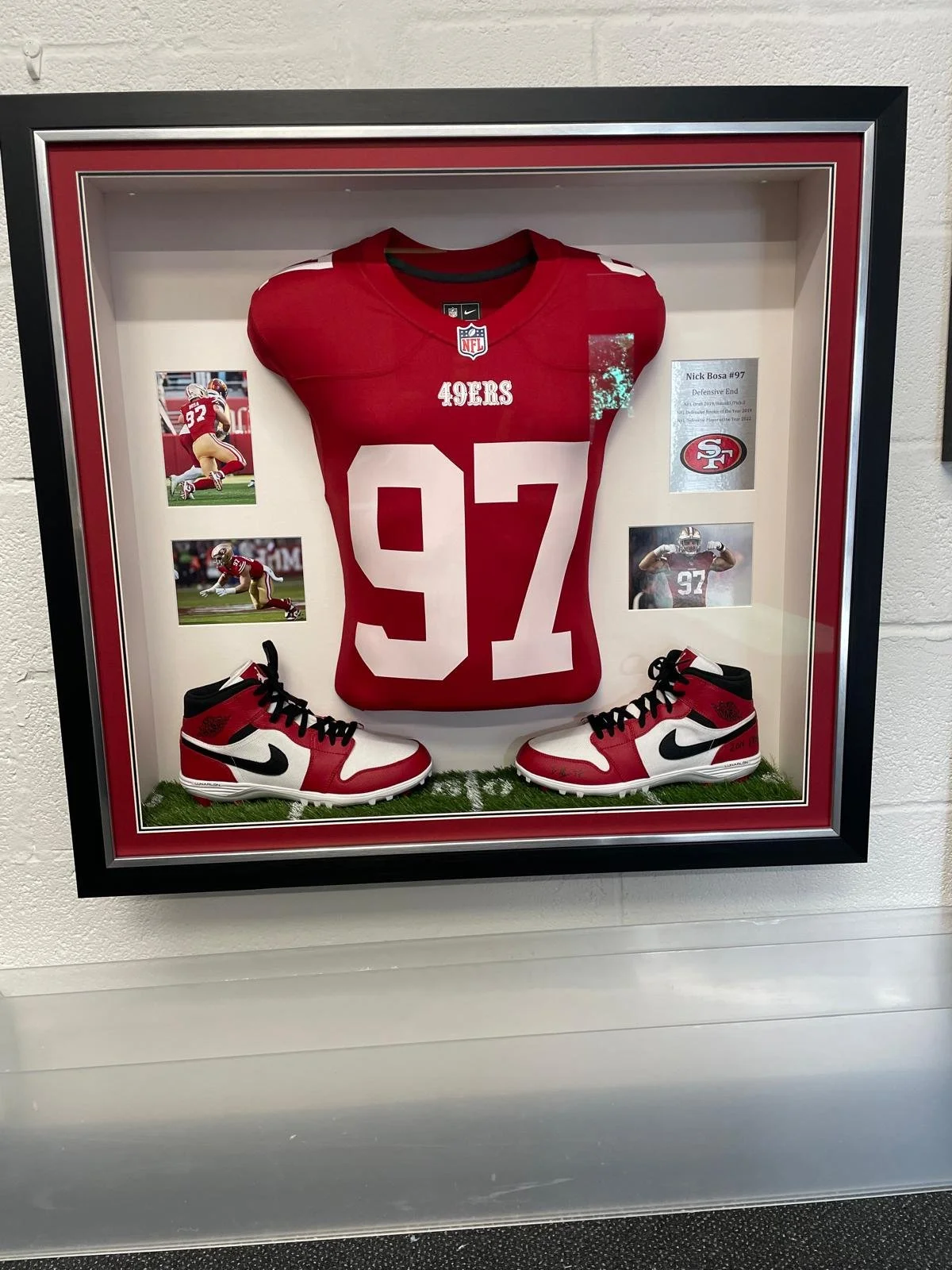 A framed display of a San Francisco 49ers football jersey numbered 97, with matching red, black, and white Nike cleats, photos of a player in action, and a small NFL note card inside a glass case.