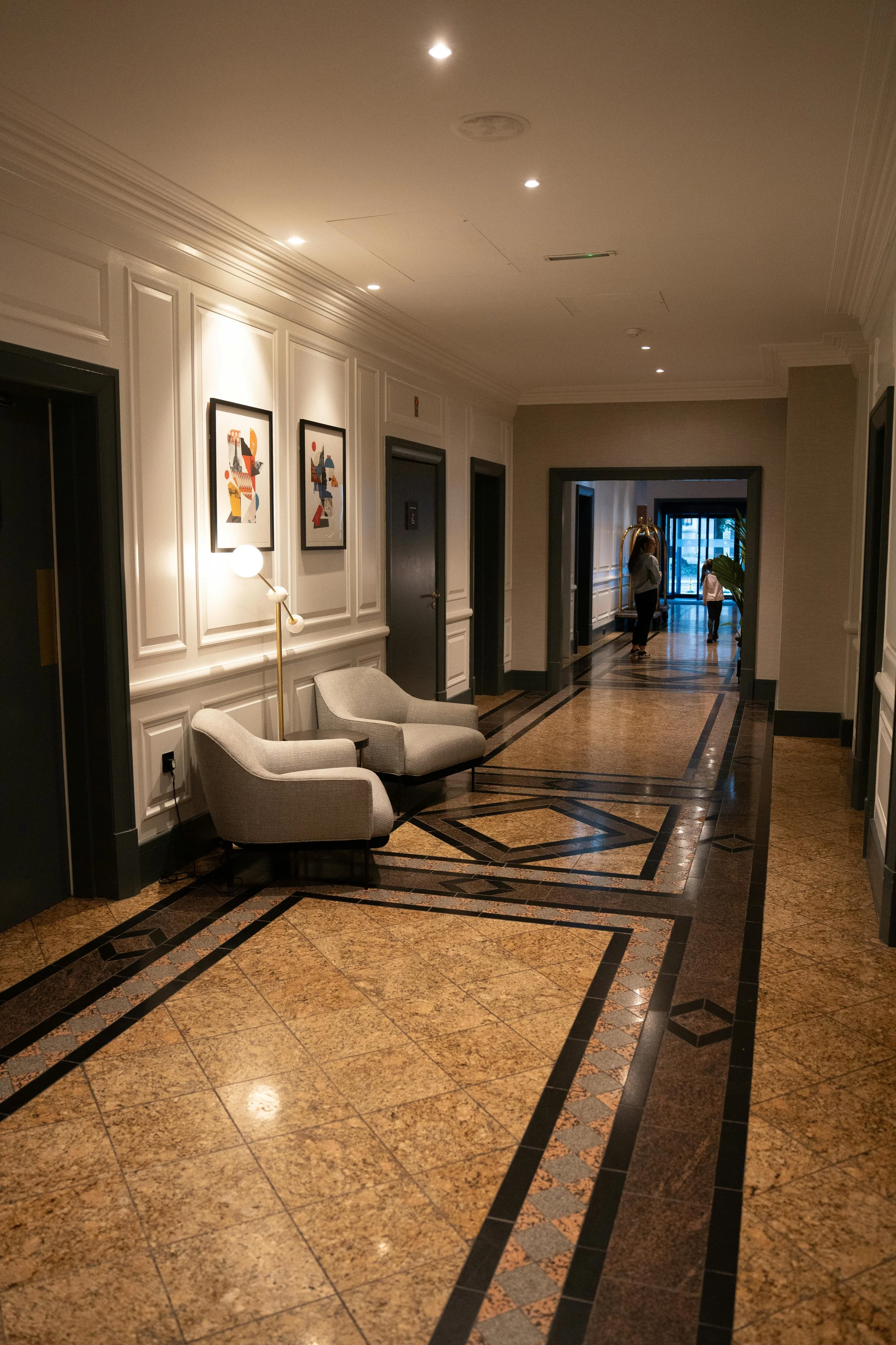 Elegant hotel hallway with patterned tiled floors, white wainscoting walls, artwork, two beige armchairs, and a floor lamp. People can be seen walking in the distance.