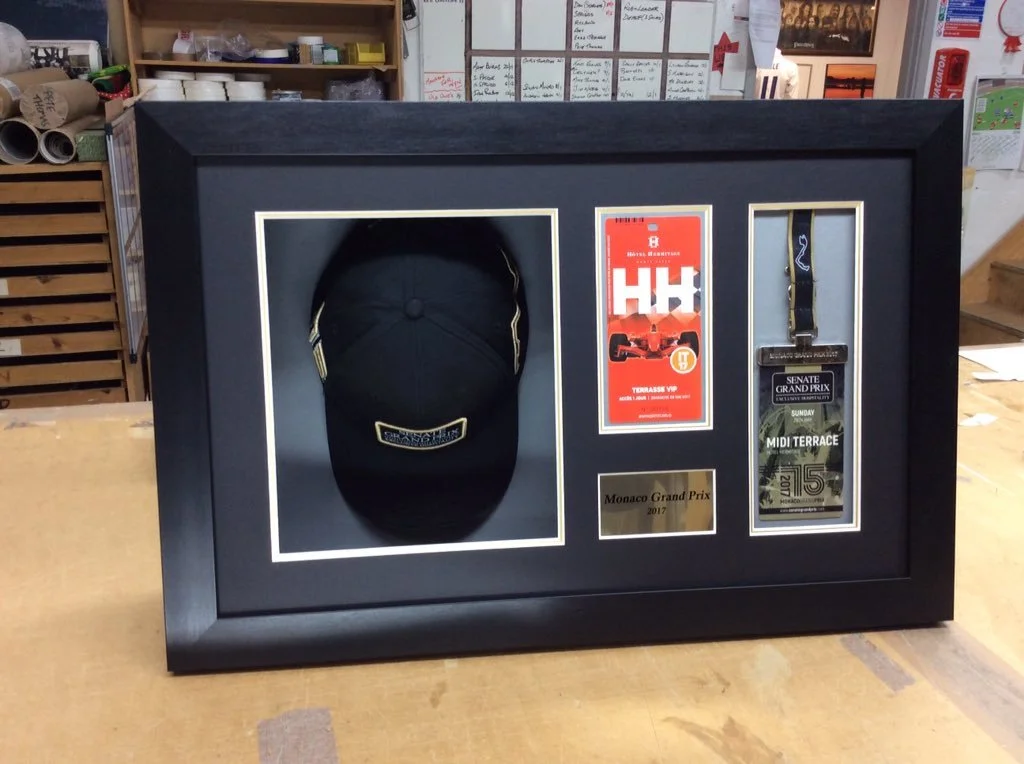 A display frame containing a black cap, two tickets, a medal, and a plaque, related to the 2017 Monaco Grand Prix, on a wooden table in a workshop