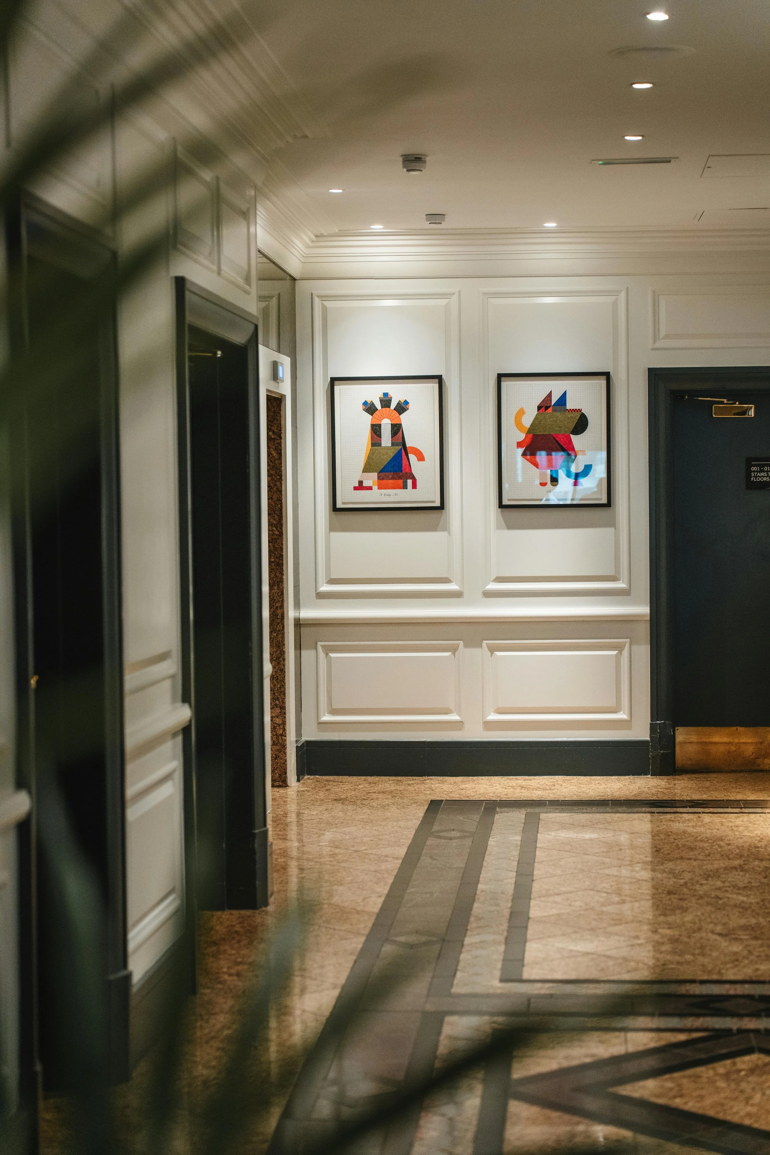 Two framed abstract artwork pieces hanging on a white wall in a hotel lobby or hallway, with decorative molding and ceiling lights.