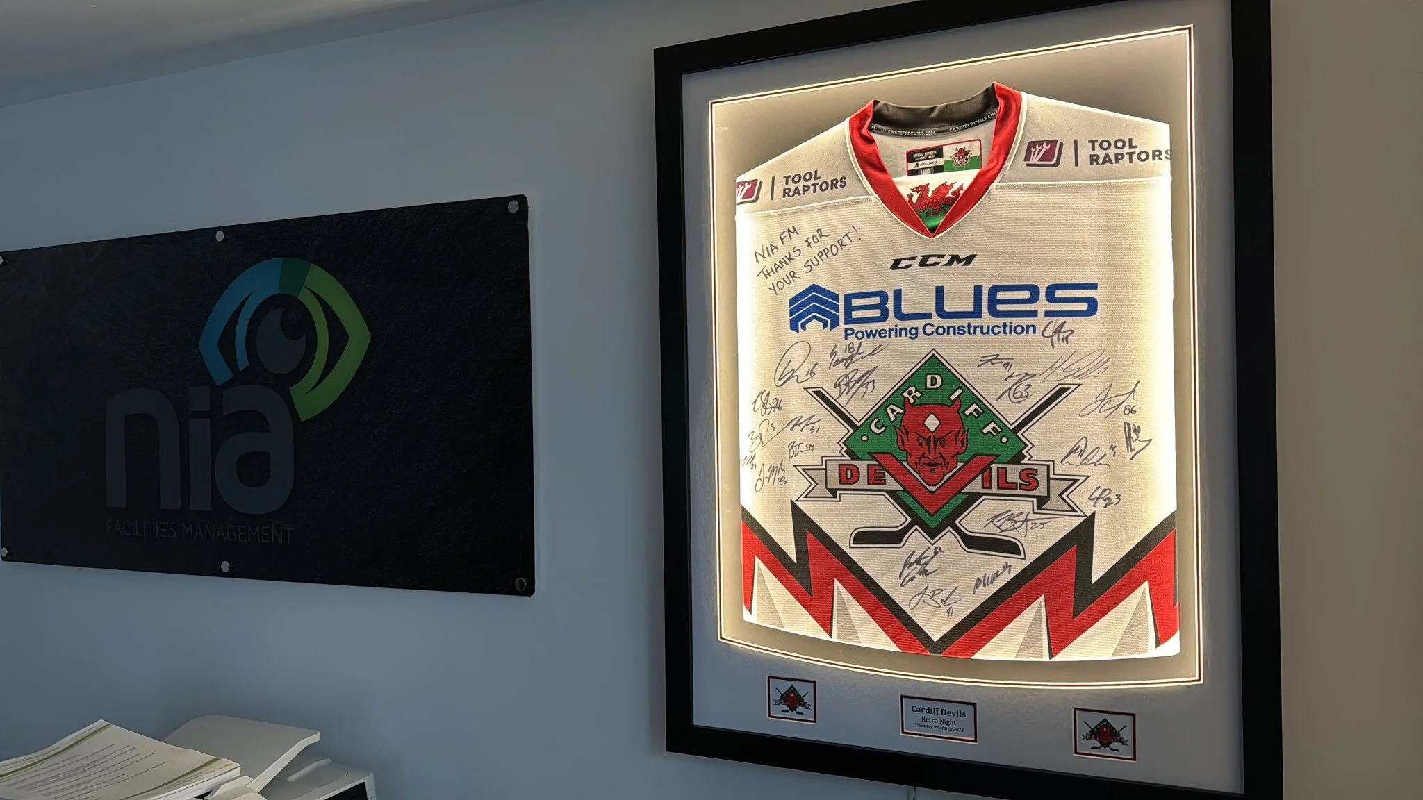 Backlit framed Cardiff Devils hockey jersey with signatures, logo, and sponsor logos, hanging on a wall in an office or sports facility.