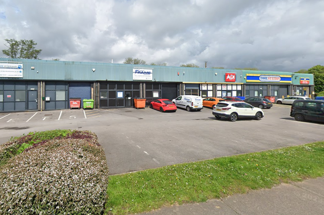 Parking lot in front of a building with several signs, including 'FAAME', 'AD', and 'TOOL STATION', with multiple parked cars and a landscaped area with bushes and grass.