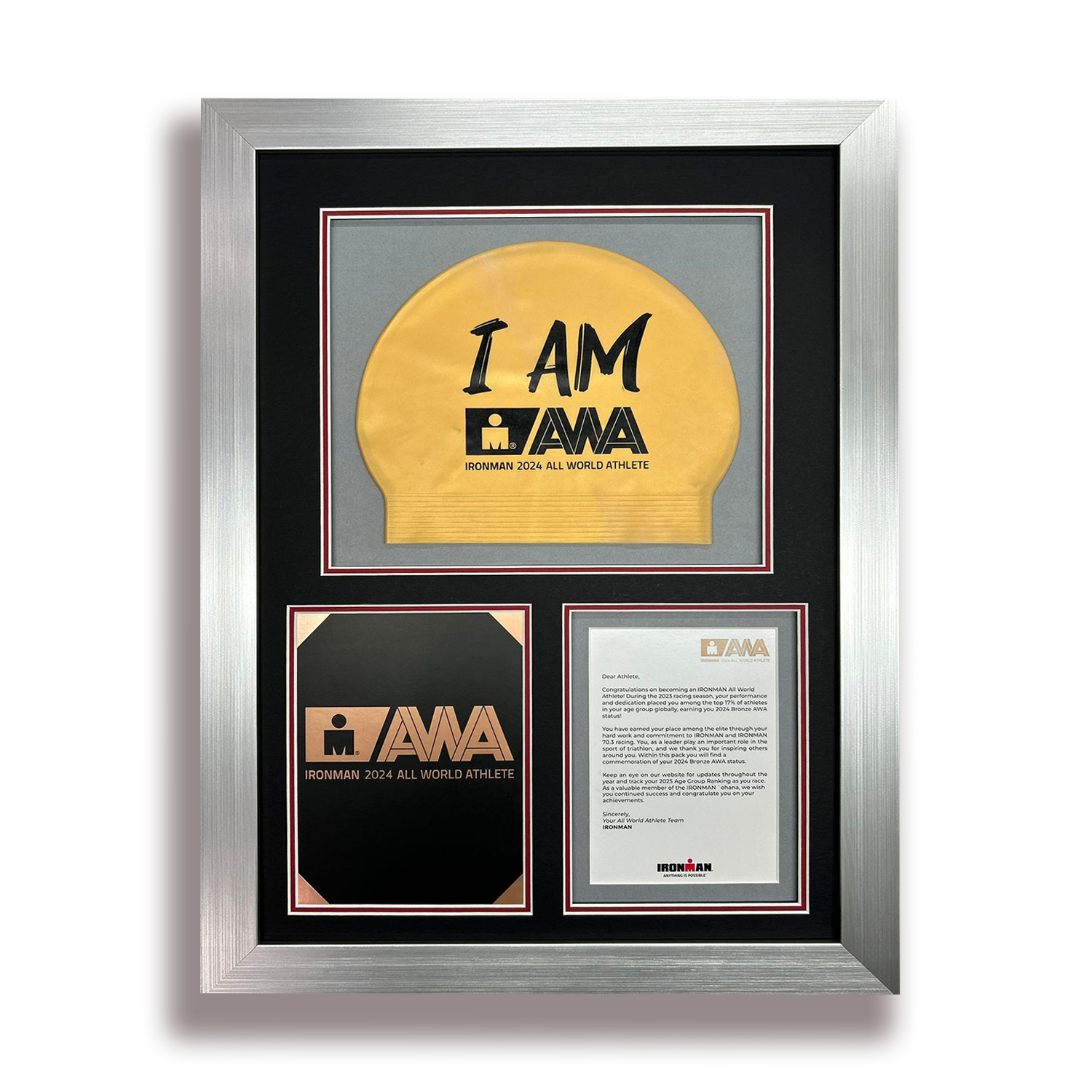Framed display of awards and recognition for an IRONMAN 2024 All World Athlete, including a gold medal, a black and copper plaque, and a letter of congratulations.