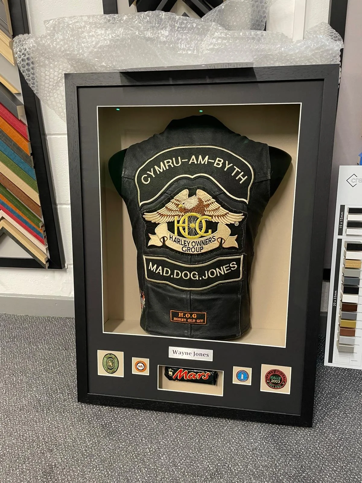 Framed display case containing a black leather vest with embroidered patches, surrounded by various stickers and labels, including a name tag that reads 'Wayne Jones'.