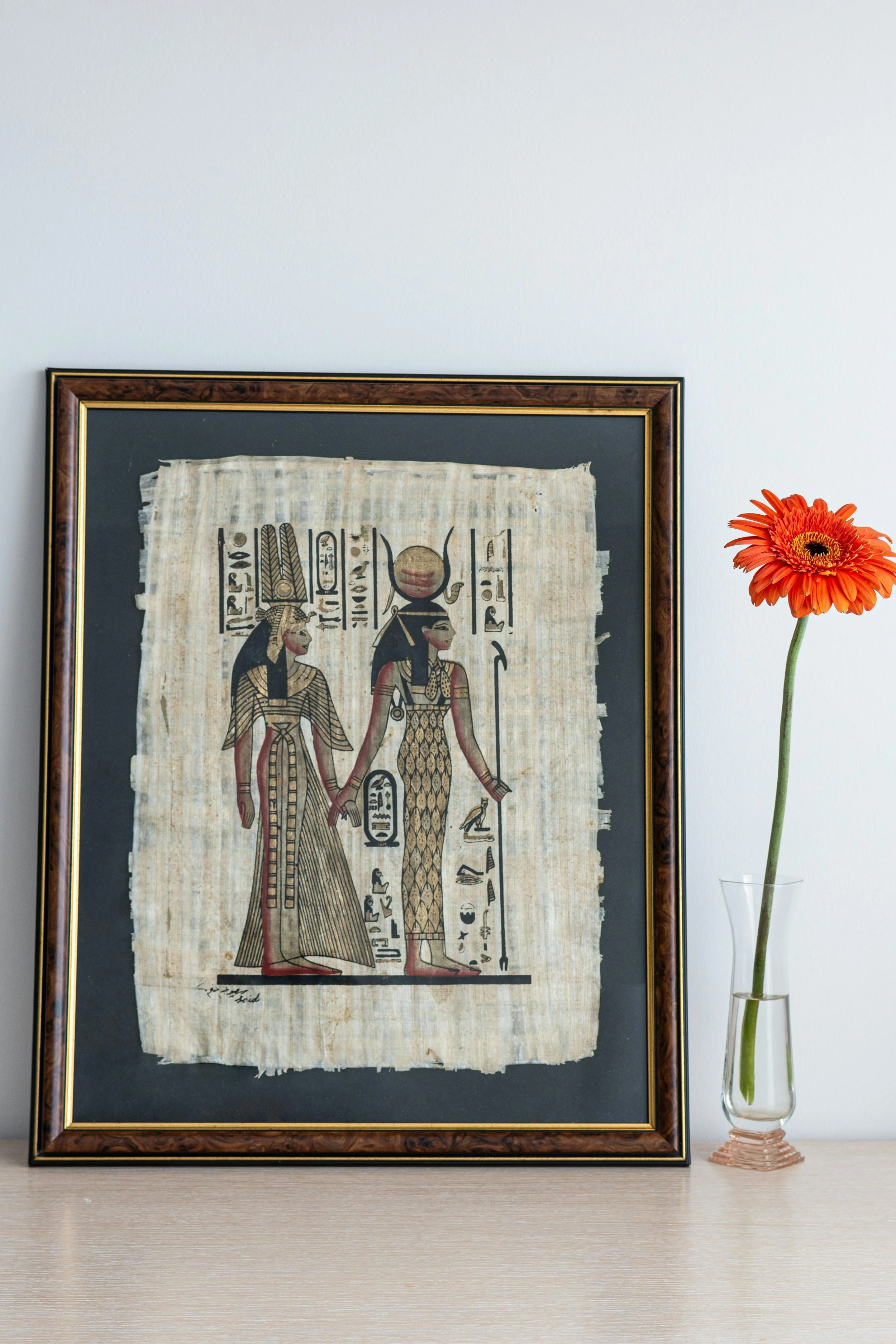 Framed ancient Egyptian artwork of two women with hieroglyphics, placed next to a small vase with a single orange gerbera daisy.