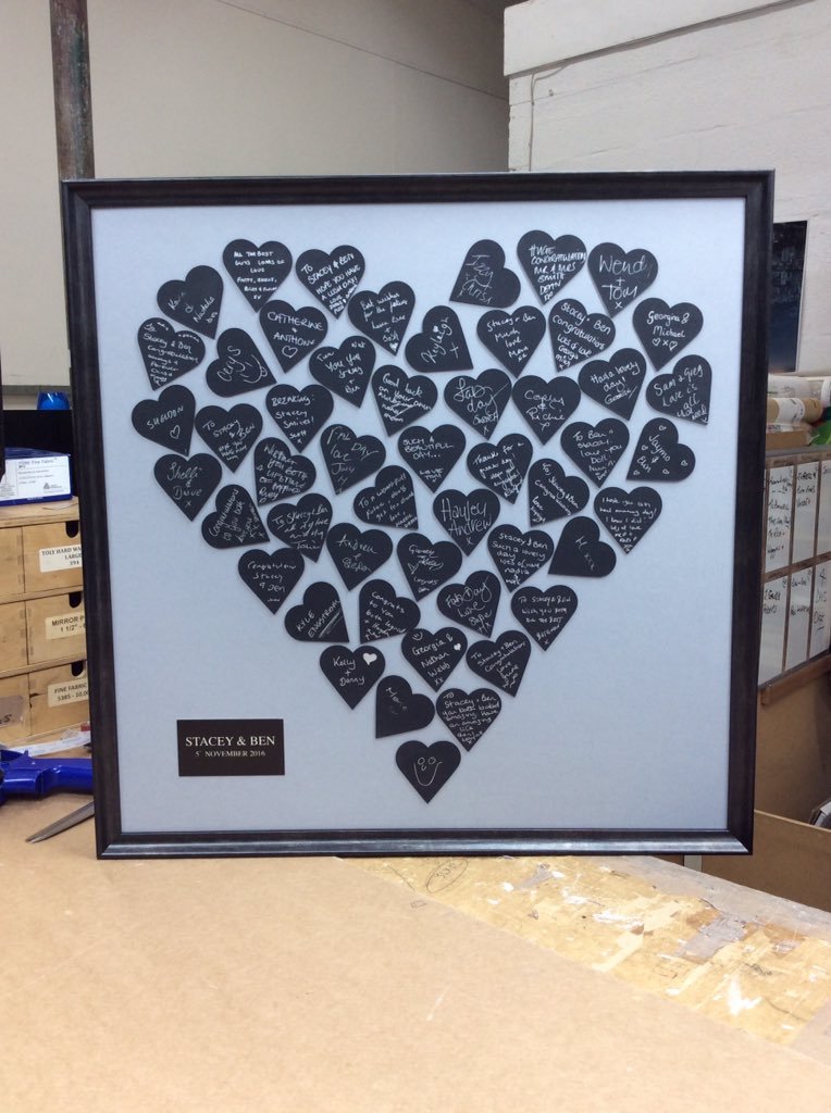 A framed display of numerous black heart-shaped notes with handwritten messages inside a larger heart shape. The notes are filled with names, well wishes, and congratulatory messages. A label at the bottom left reads 'Stacey & Ben 5 November 2016'.
