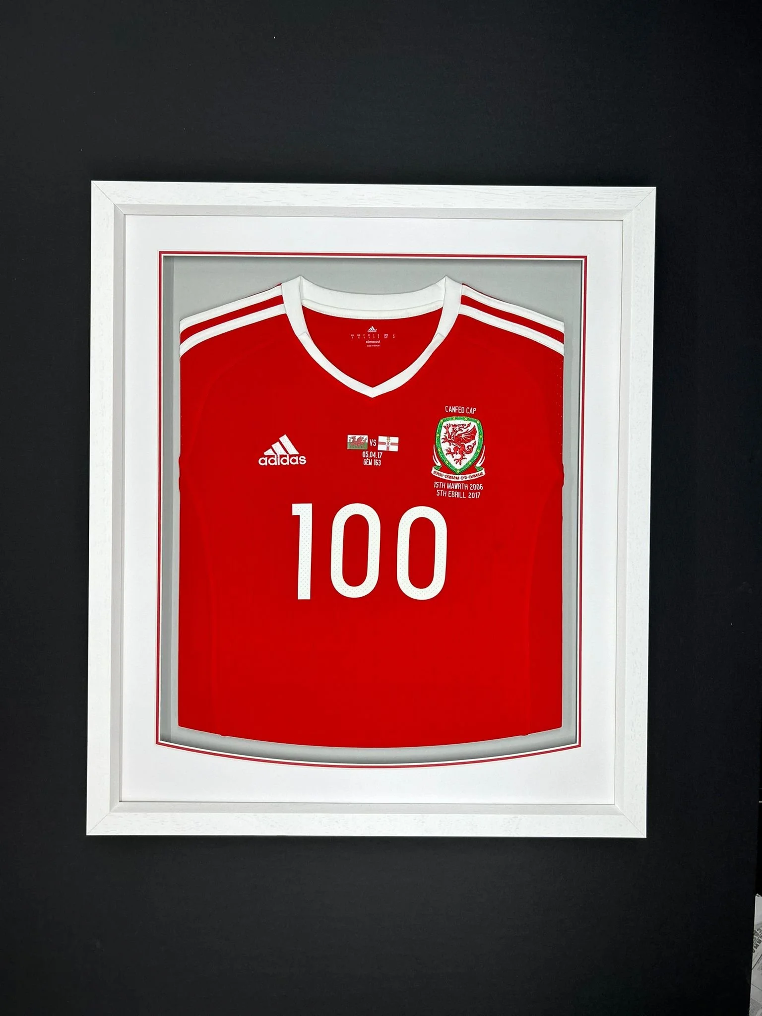 Framed Welsh football jersey with the number 100, white frame, black background.