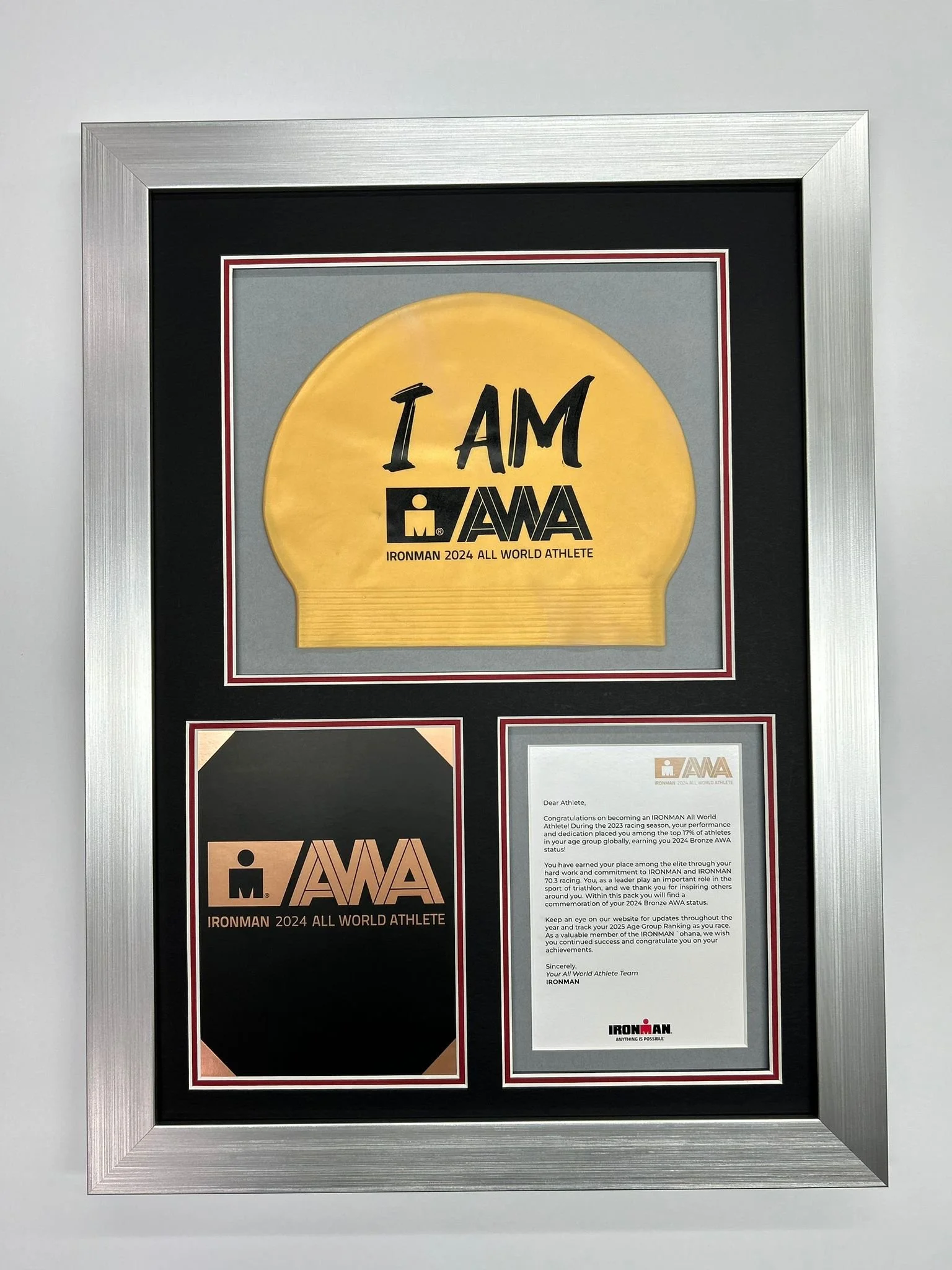 Framed display celebrating achievement in the 2024 IRONMAN All World Athlete program, including a yellow medal with the words "I AM" and the IRONMAN logo, and a certificate or letter from IRONMAN recognizing the athlete's accomplishment.