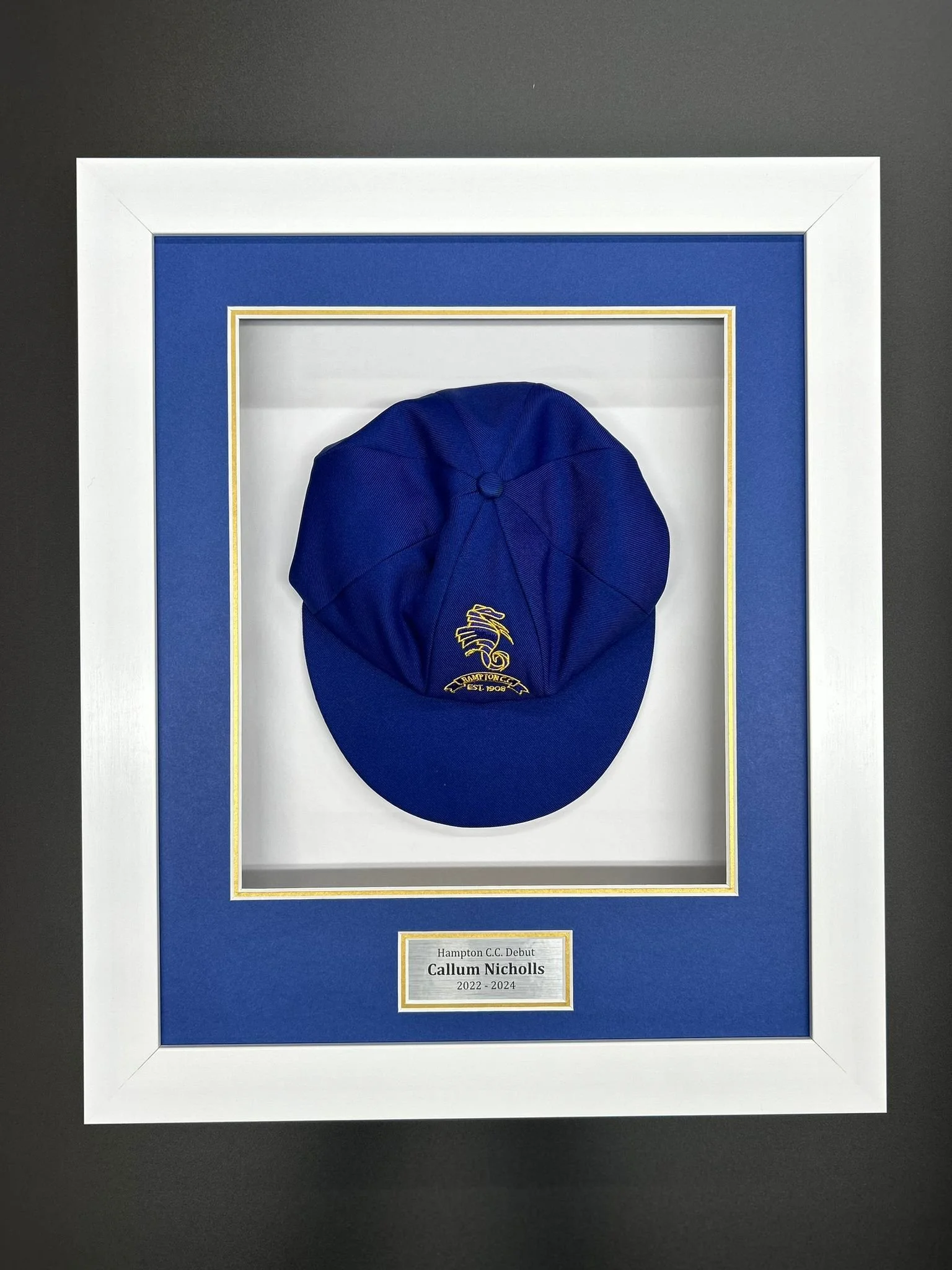 A framed blue cap with a yellow logo, commemorating Callum Nicholls' debut for Hampton C.C. from 2022 to 2024.