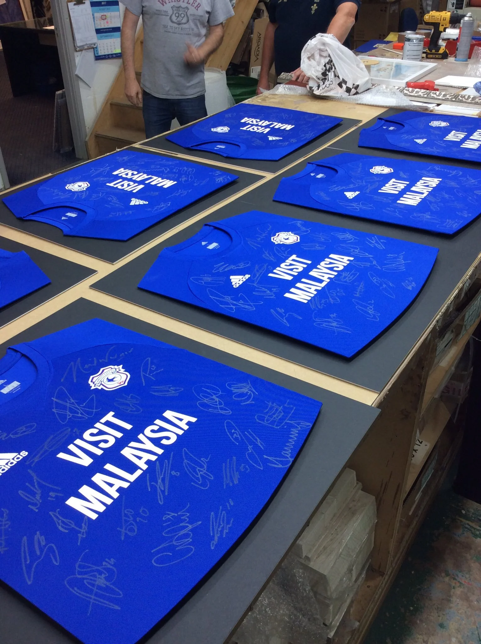 Blue sports jerseys with 'Visit Malaysia' printed on them, signed by multiple individuals, laid out on a worktable for signing or autographs.