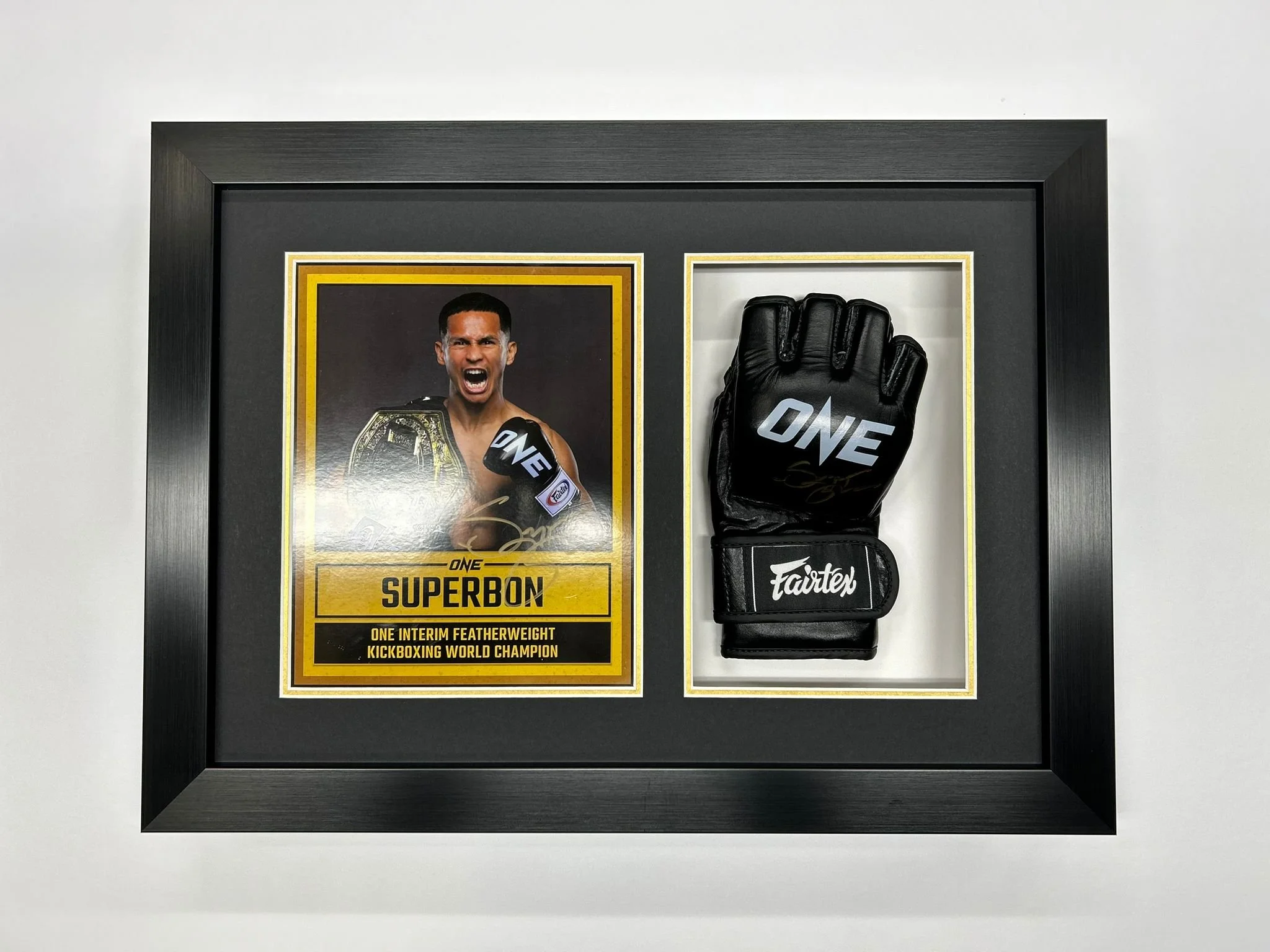 Framed display of a kickboxing champion, showing a photo of a man with a championship belt and a glove with the ONE logo, along with his autograph and title 'Superbon, One Interim Featherweight Kickboxing World Champion'.