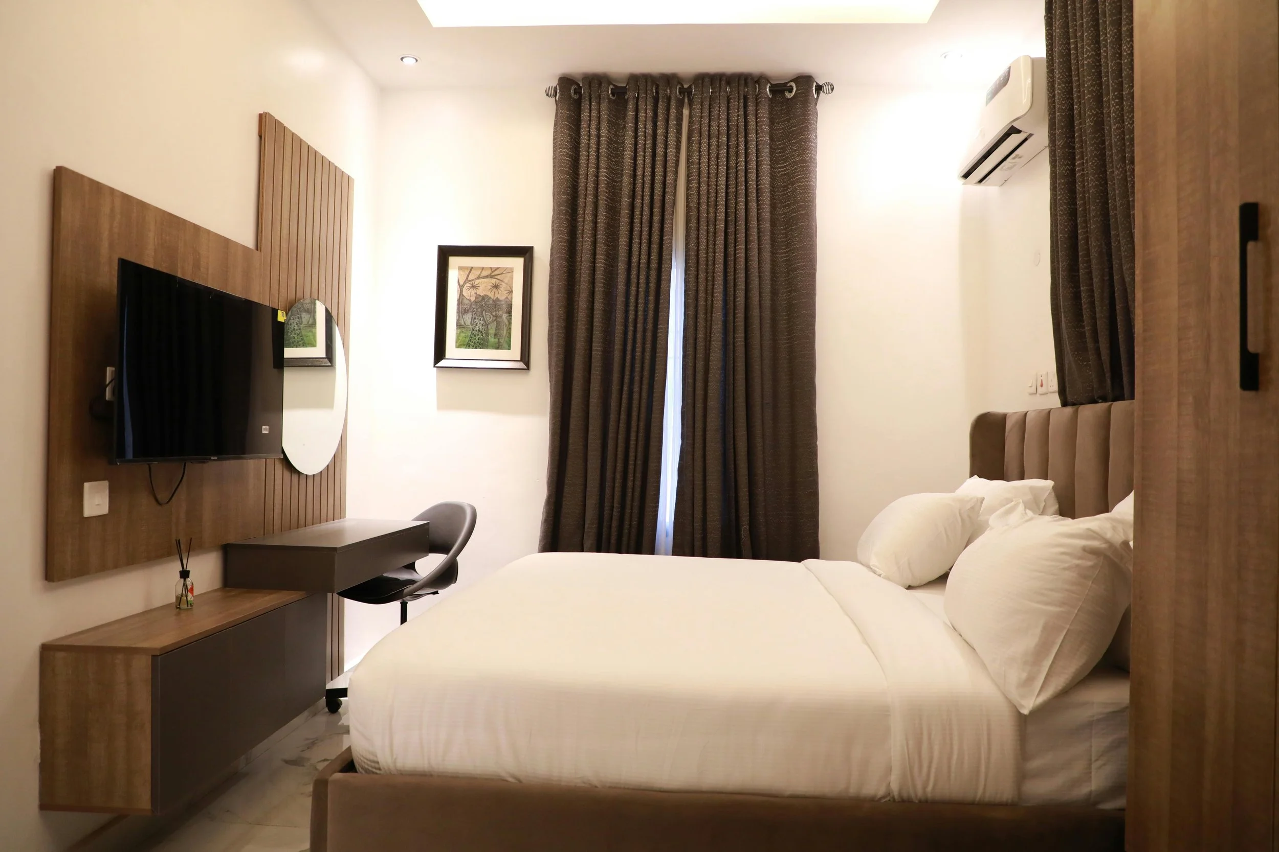 Hotel room with a bed, brown curtains, wall art, wall-mounted TV, desk, chair, and air conditioning unit.