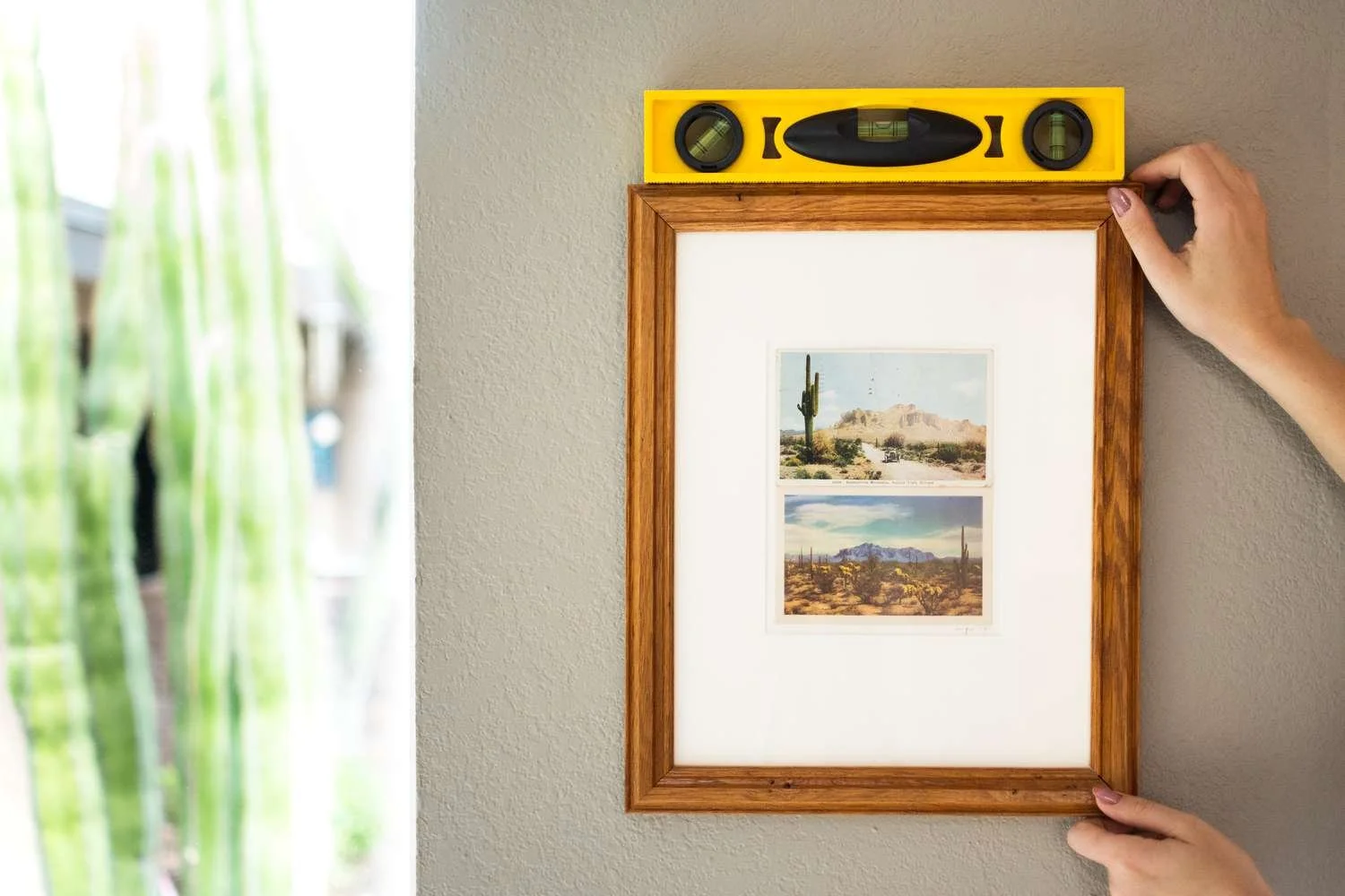Person using a level tool to hang a wooden framed picture of desert landscapes on a wall.