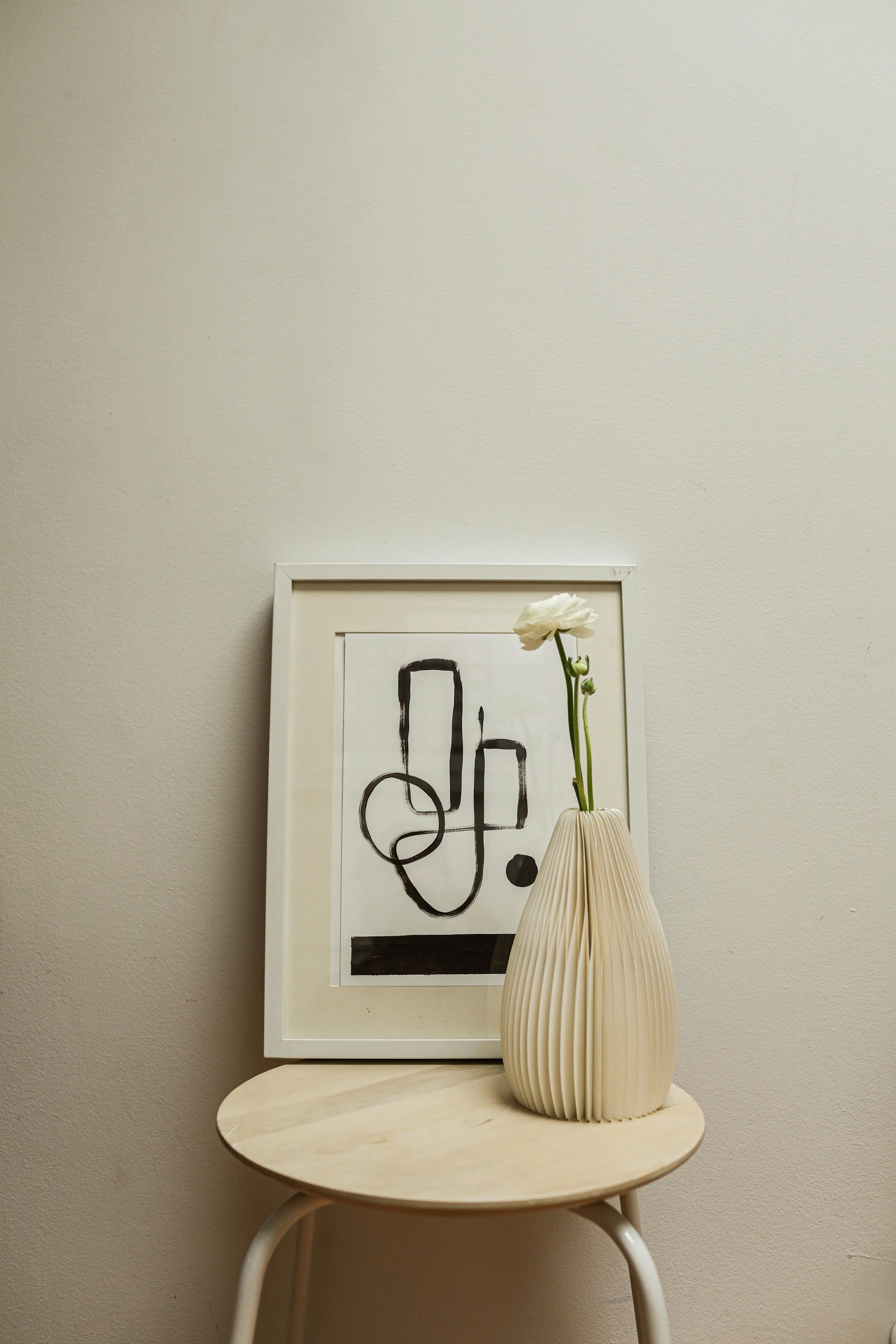 A white framed abstract black and white artwork rests on a small round beige table, with a white pleated vase and a single white flower beside it.