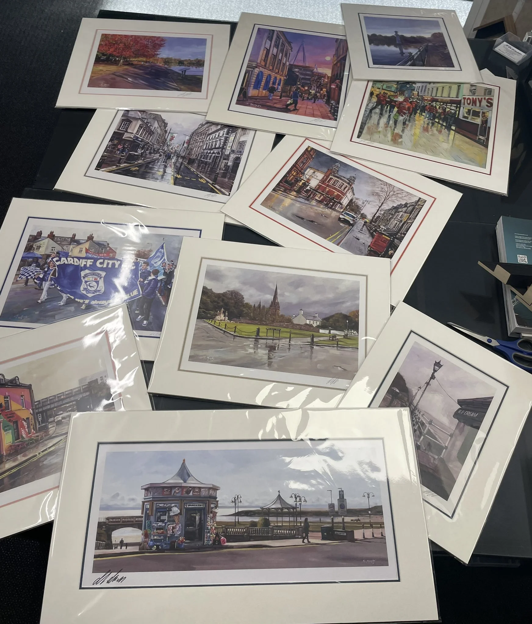 Collection of framed landscape and cityscape paintings, including scenes of streets, a park, a pier, and a parade, displayed on a table.