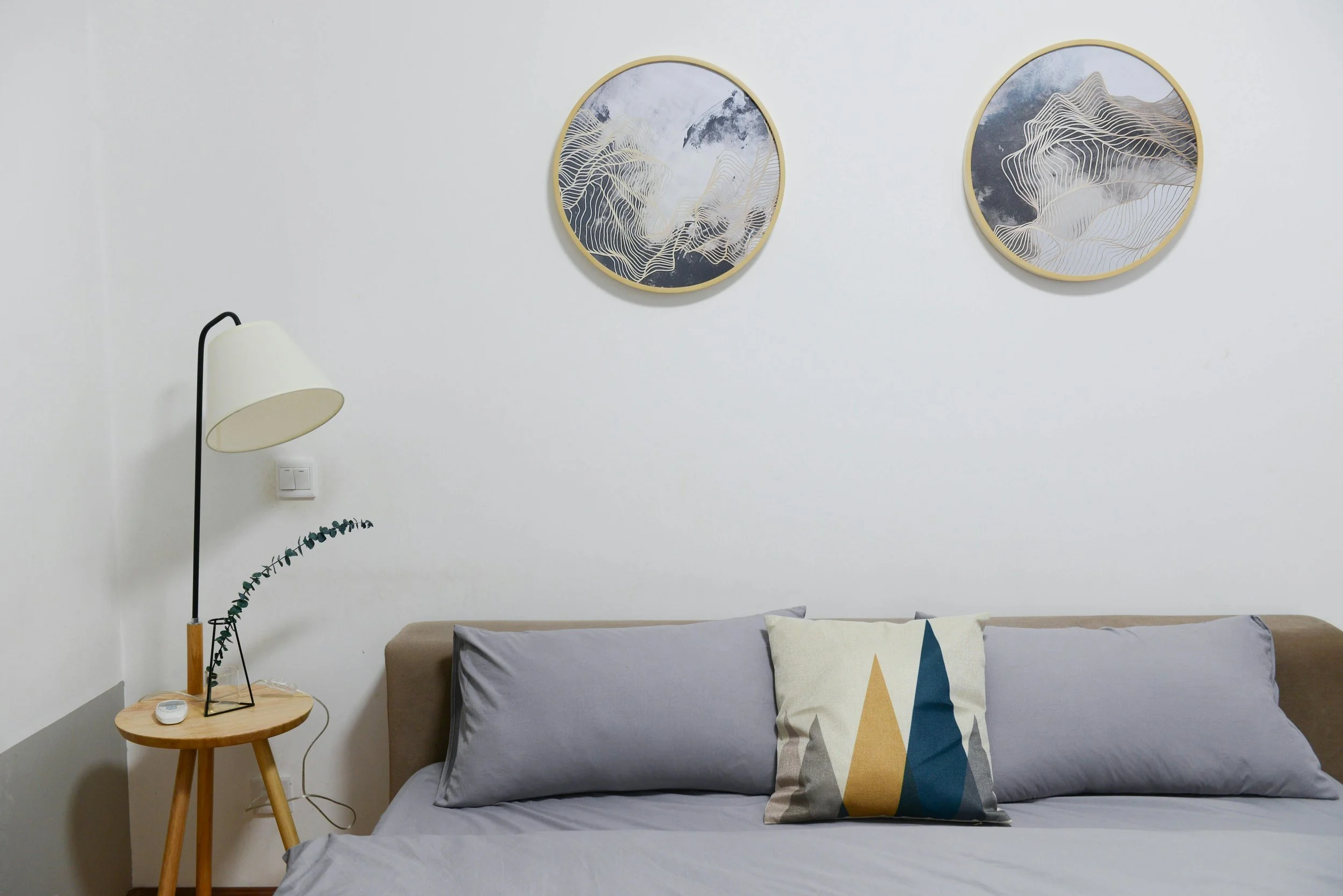 A bedroom with a beige bed, a gray pillow with a geometric design, two decorative wall art pieces with abstract mountain patterns, a wooden bedside table with a white lamp, a small plant, and a circular device.