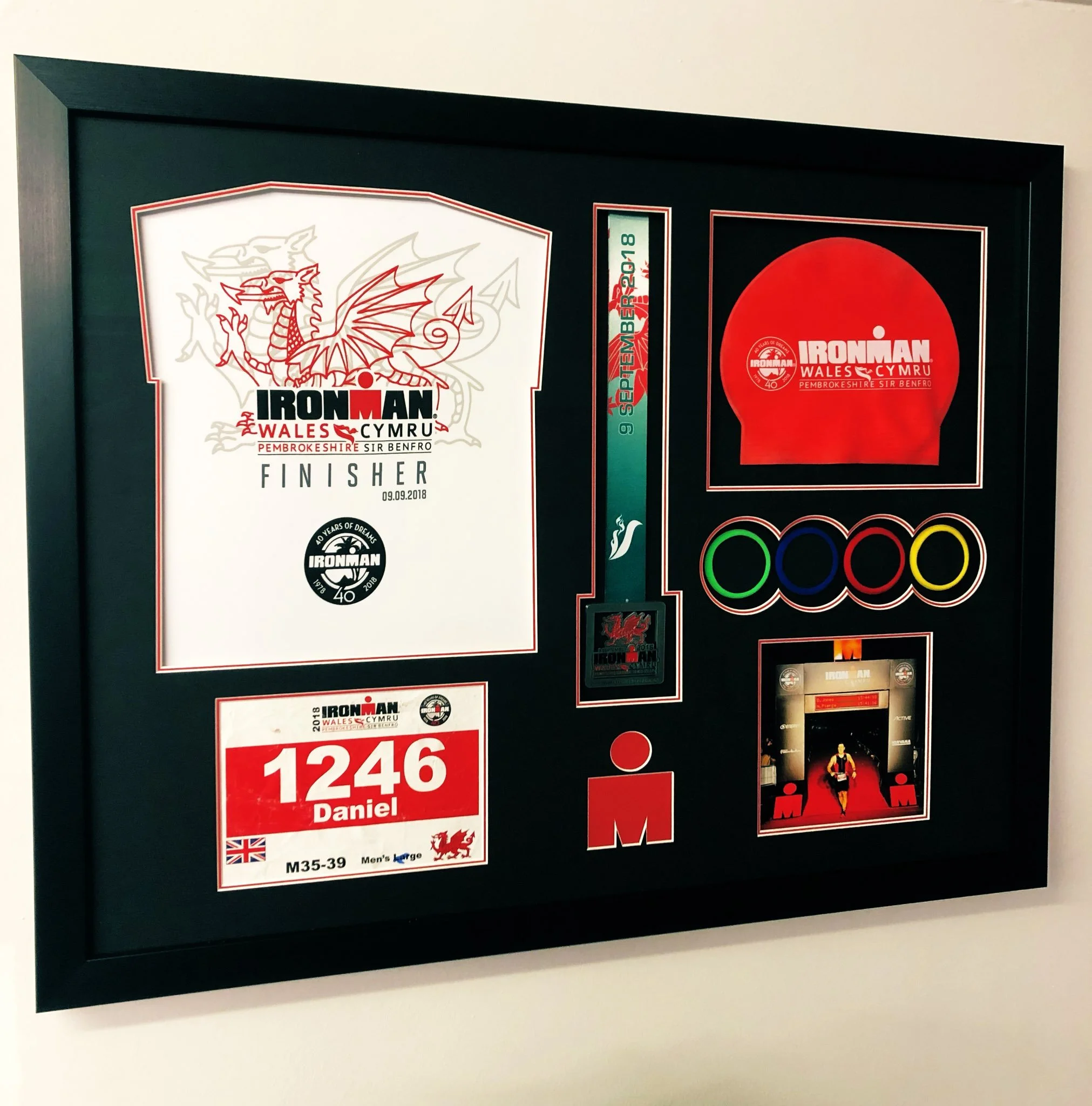 Frame with Ironman finisher memorabilia including a race bib, medal, shirt, and photos from the 2018 Wales Ironman event.