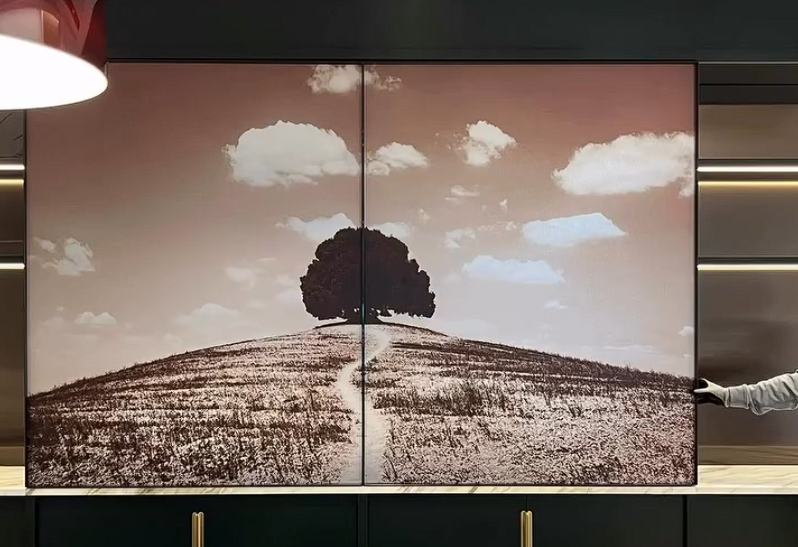 A large wall mural depicts a landscape with a single tree on a hill, under a sky with scattered clouds. The mural is divided into two panels. A person is partially visible on the right, holding the edge of the mural.