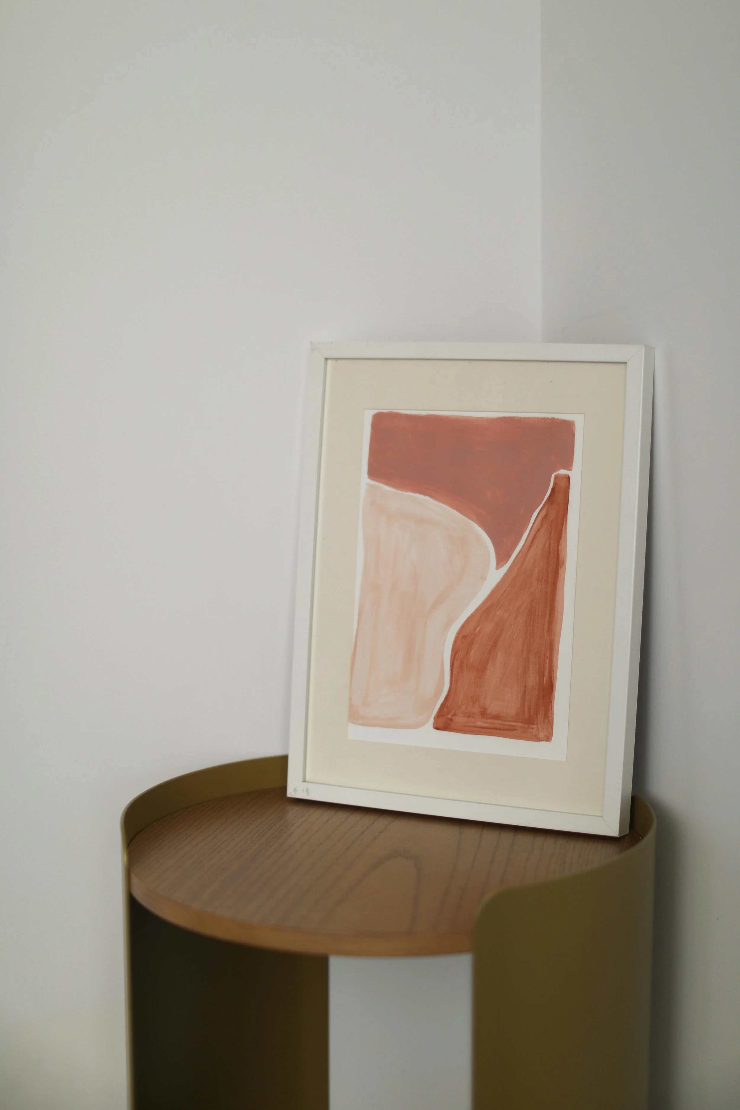Framed abstract art with earthy tones of brown, beige, and terracotta, resting on a round wooden side table with a gold-colored rim against a plain white wall.