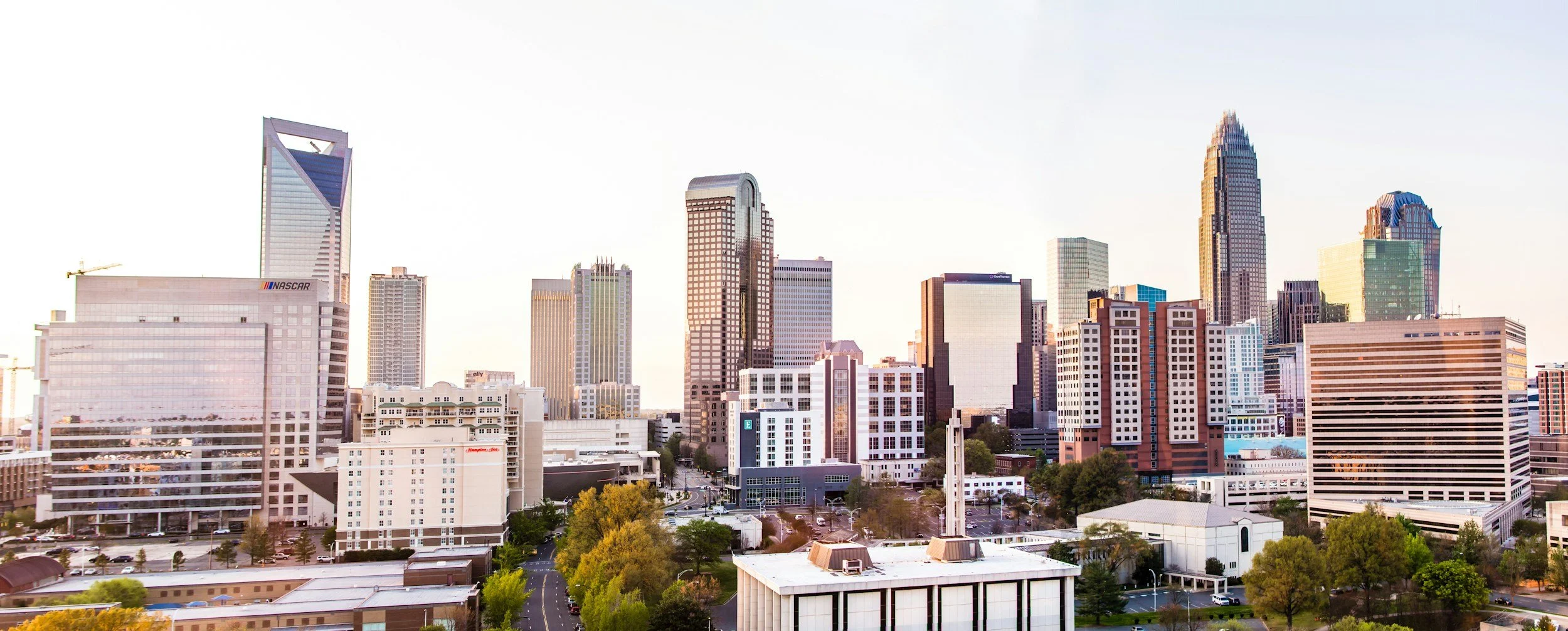 Skyline of Charlotte, North Carolina with tall buildings and skyscrapers, including the Bank of America corporate headquarters, with trees and a partly cloudy sky.