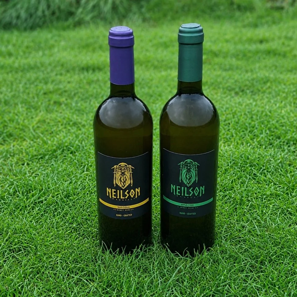 Two bottles of wine standing on grass, one with a purple cap and label, the other with a green cap and label, both labeled Neilson with a lion logo.