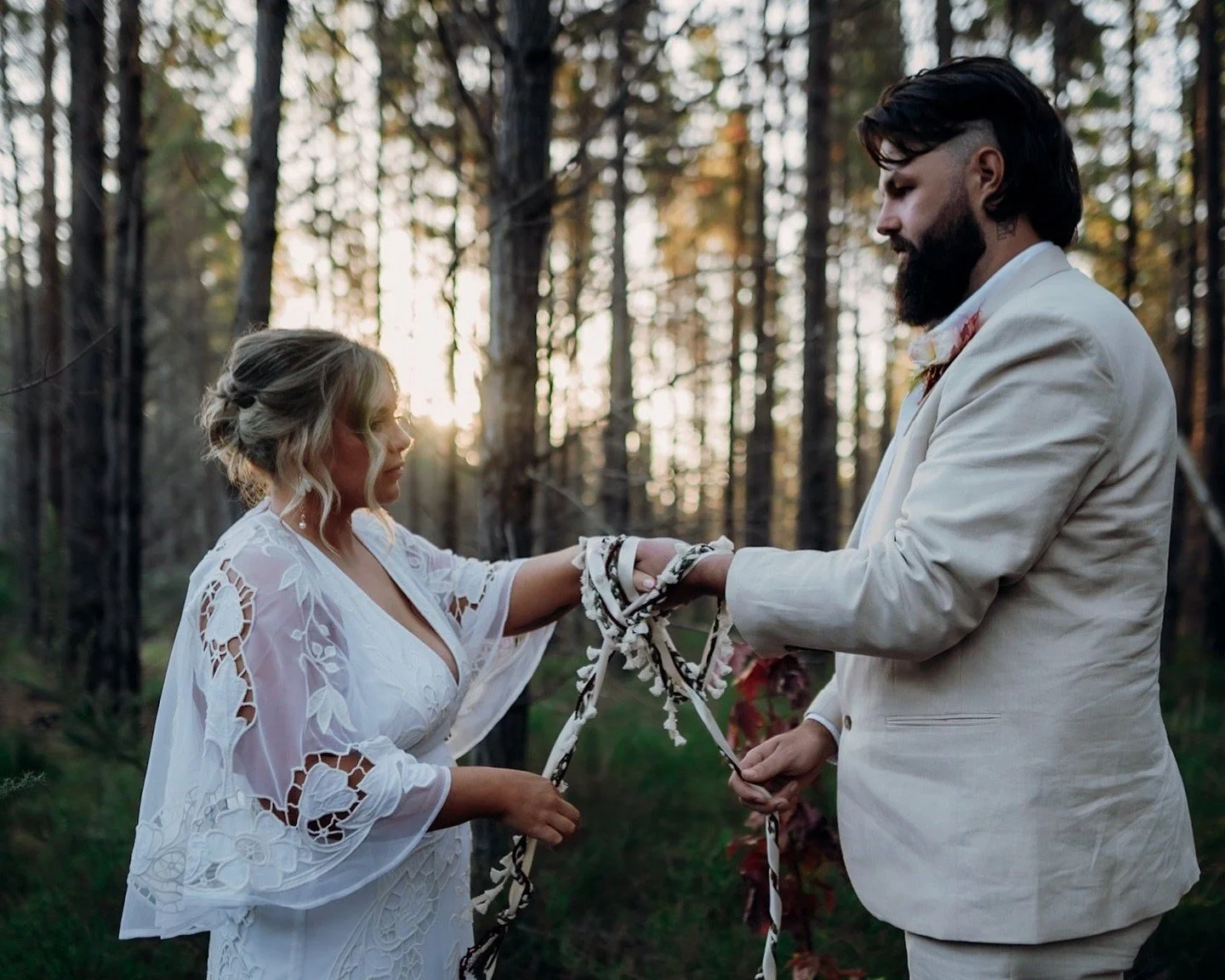 A couple exchanging vows during a wedding ceremony in a forest, with the bride in a white lace dress and the groom in a light-colored suit, holding hands amid tall trees and sunlight filtering through.