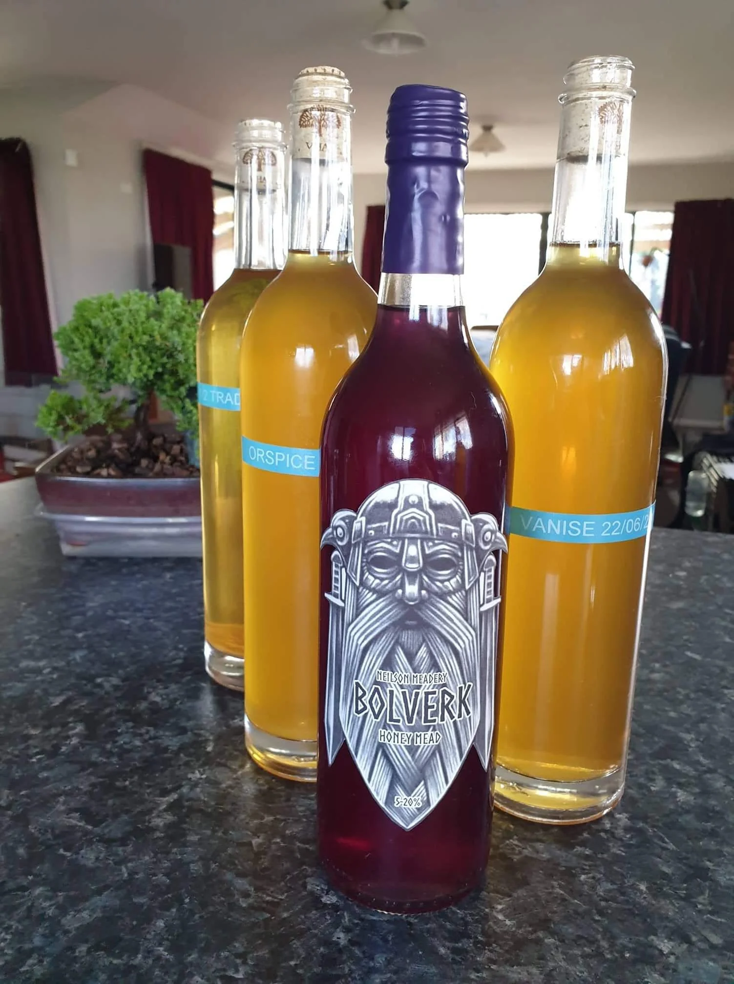 Four bottles on a kitchen counter, three containing a clear yellow liquid and one with dark red liquid, with a plant and window in the background.