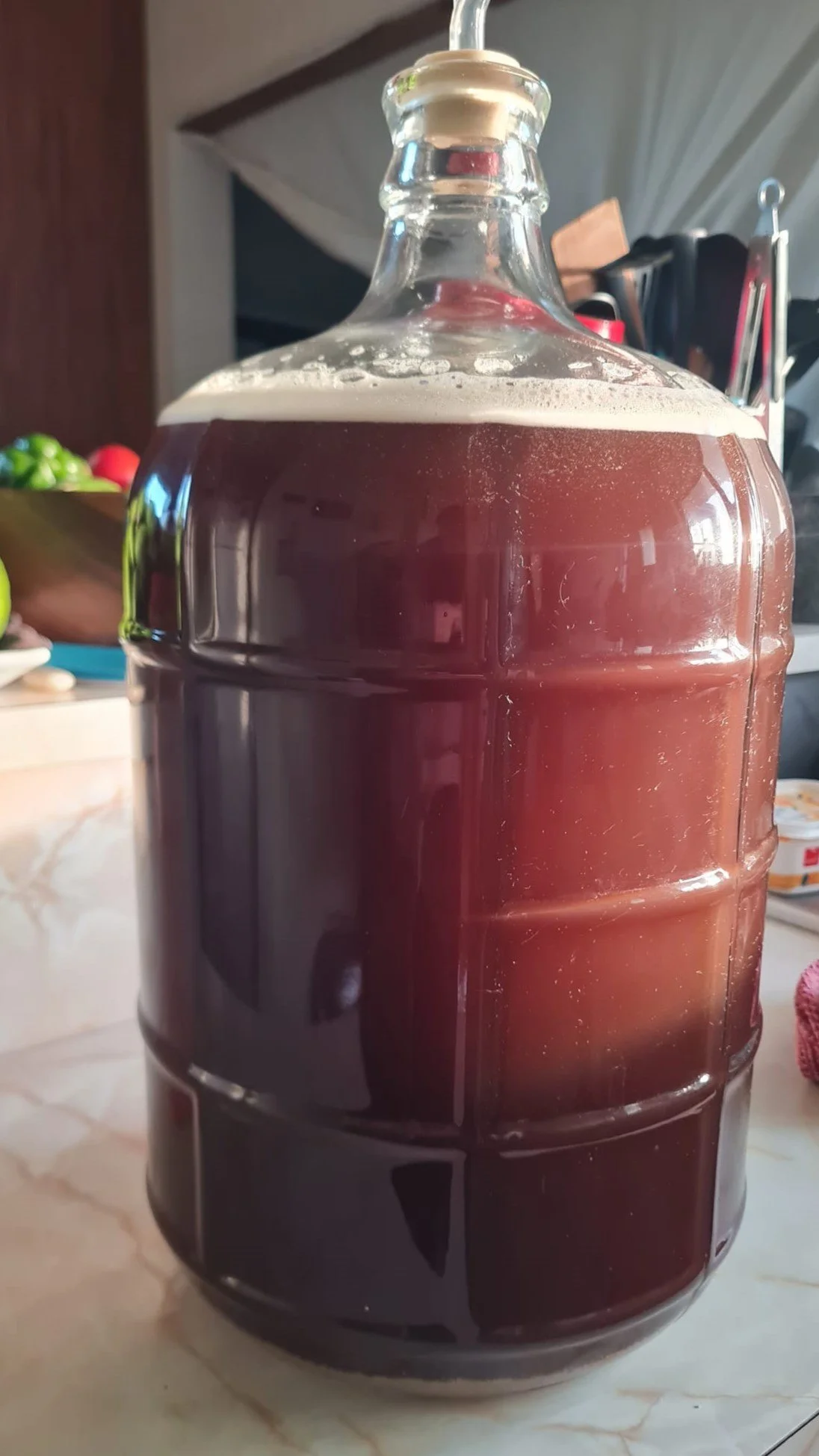 A large glass jug filled with a dark beverage, with a straw inserted through the lid, placed on a kitchen countertop.