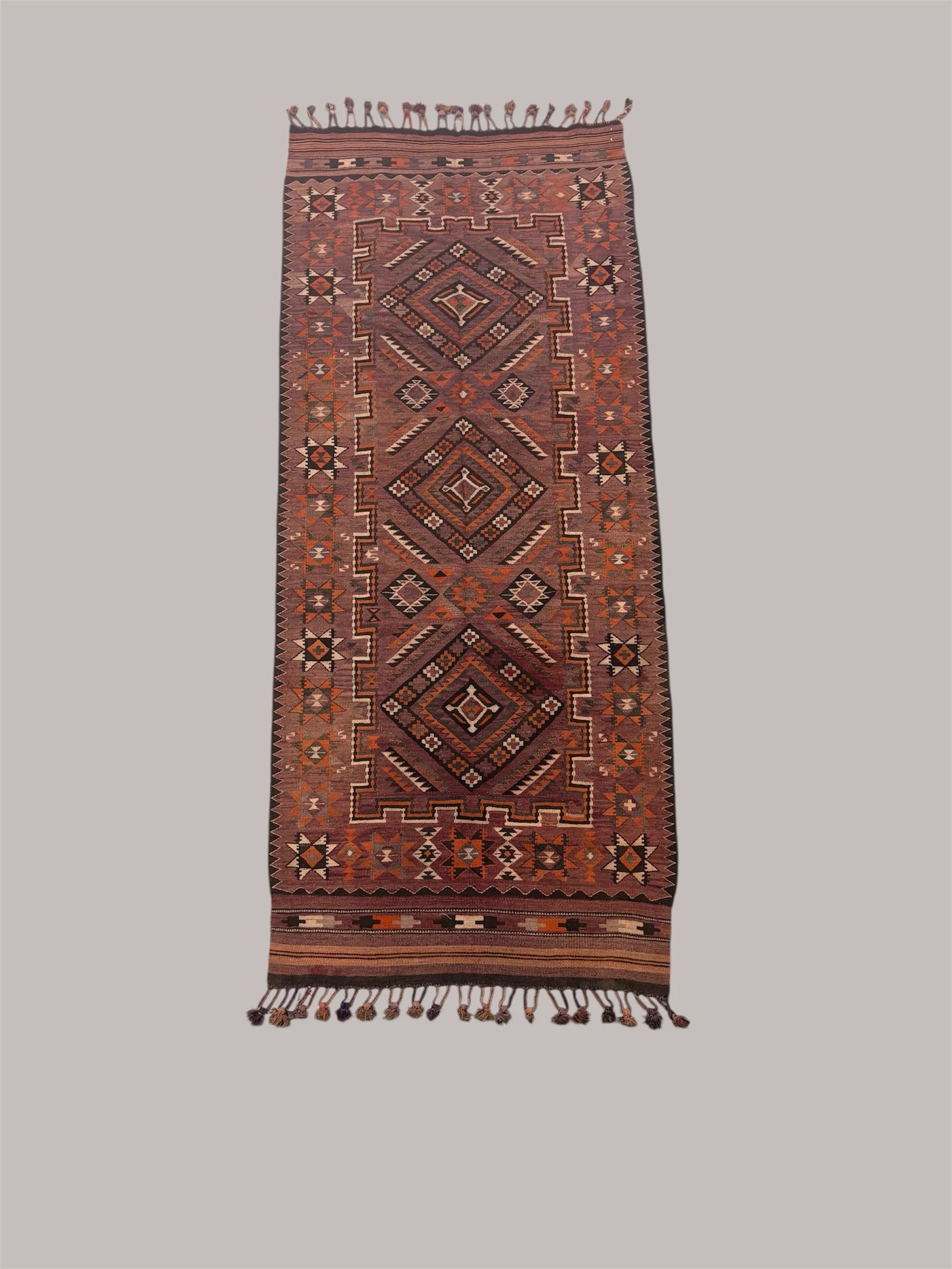 An elongated woven rug with geometric patterns in shades of red, orange, black, and cream, featuring diamonds and star motifs, and fringes at both ends.