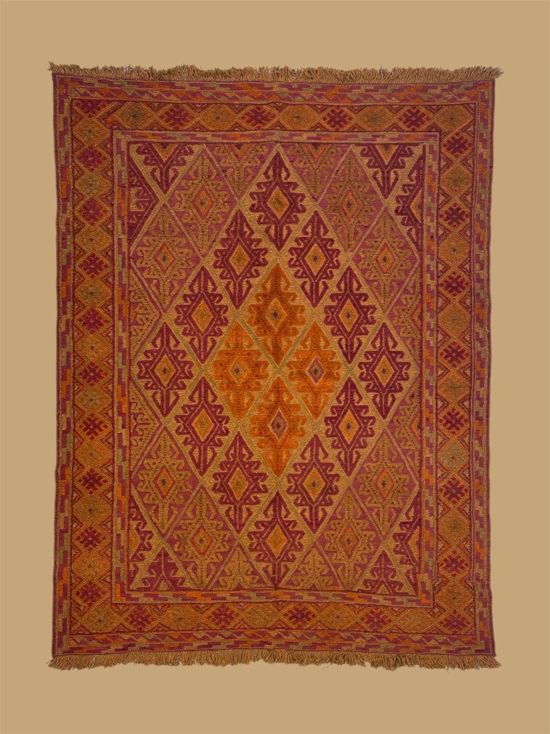 An orange and red traditional patterned woven rug with diamond and geometric designs, fringed edges, and multiple borders, hanging against a beige background.