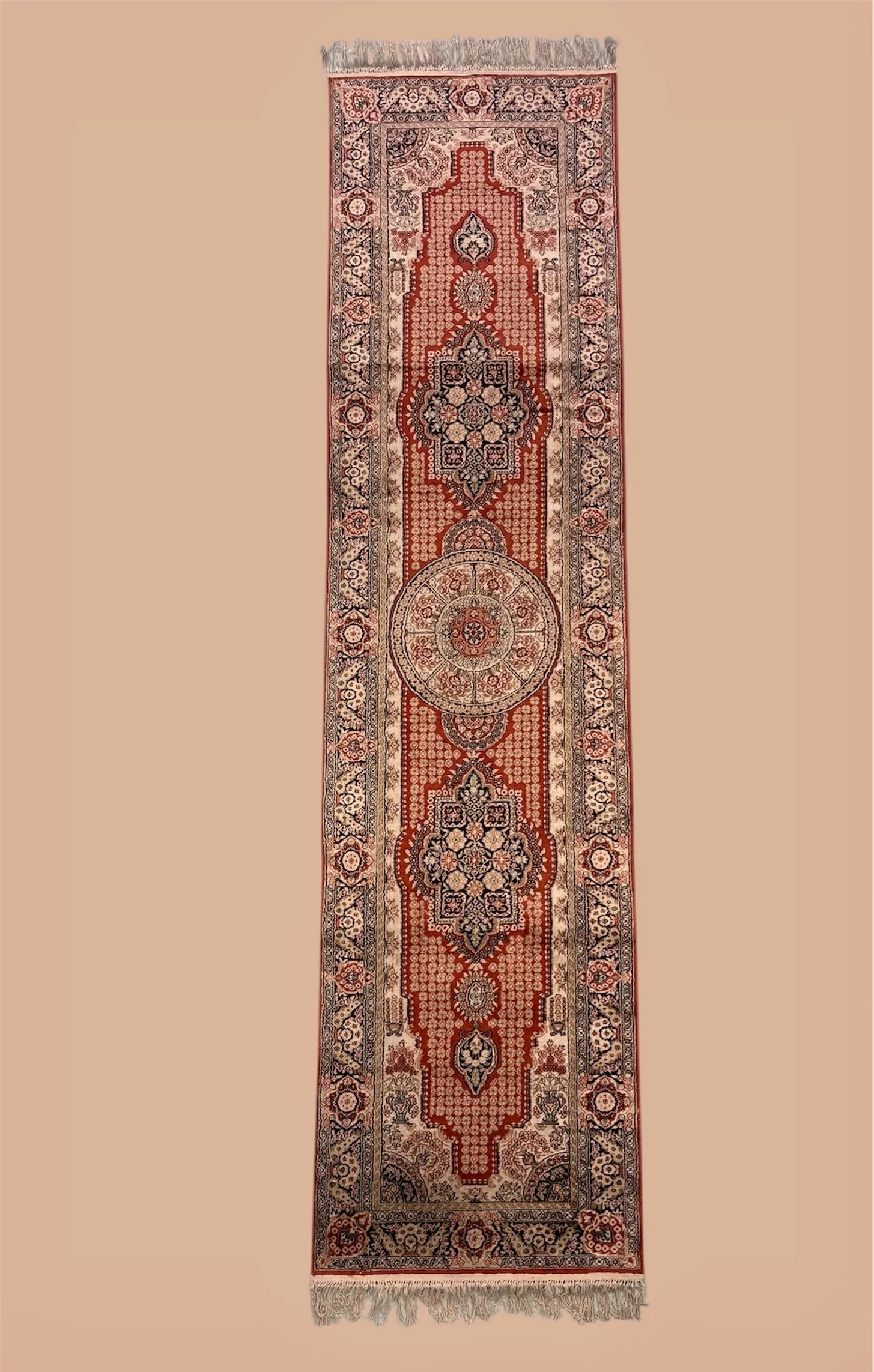 A long, narrow Persian-style rug with a central medallion design, intricate floral patterns, and a red, beige, and black color scheme, with fringes on both ends.