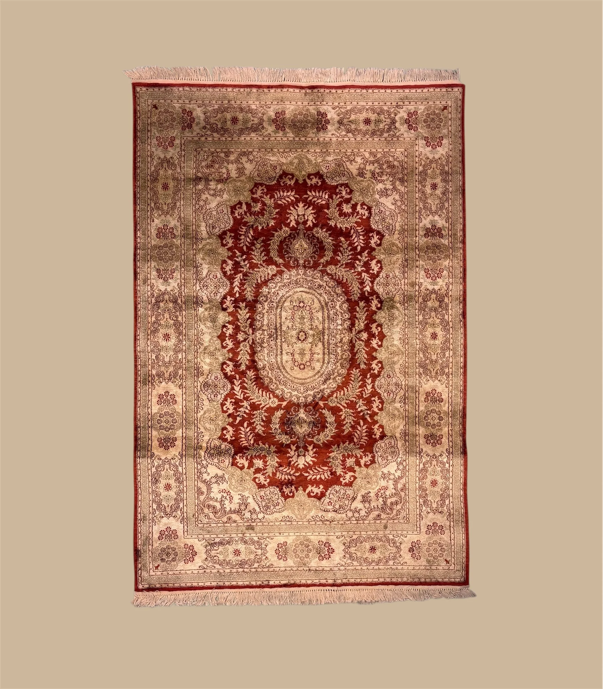 An ornate area rug with a red, beige, and brown floral and geometric pattern, fringed edges, on a plain background.