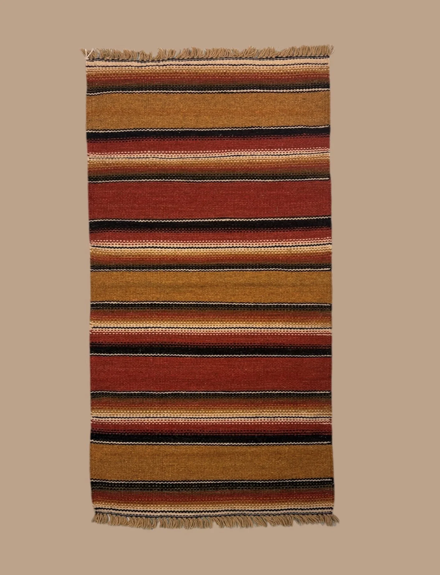 A multicolored woven rug with horizontal stripes in shades of brown, red, black, and cream, with fringed edges at the top and bottom.