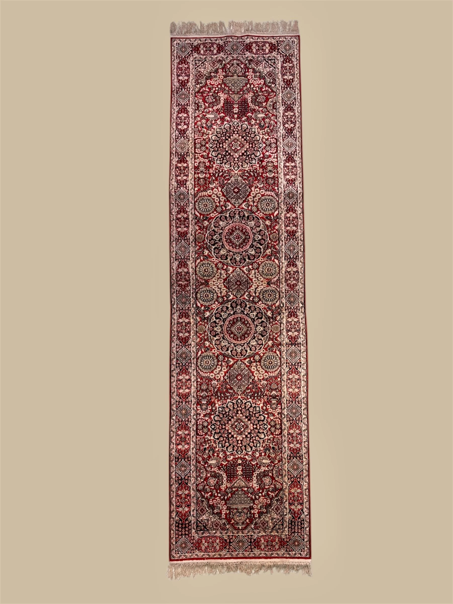 A long, narrow Persian-style rug with intricate red, black, white, and beige floral and geometric patterns, fringes at both ends, laid flat on a neutral background.