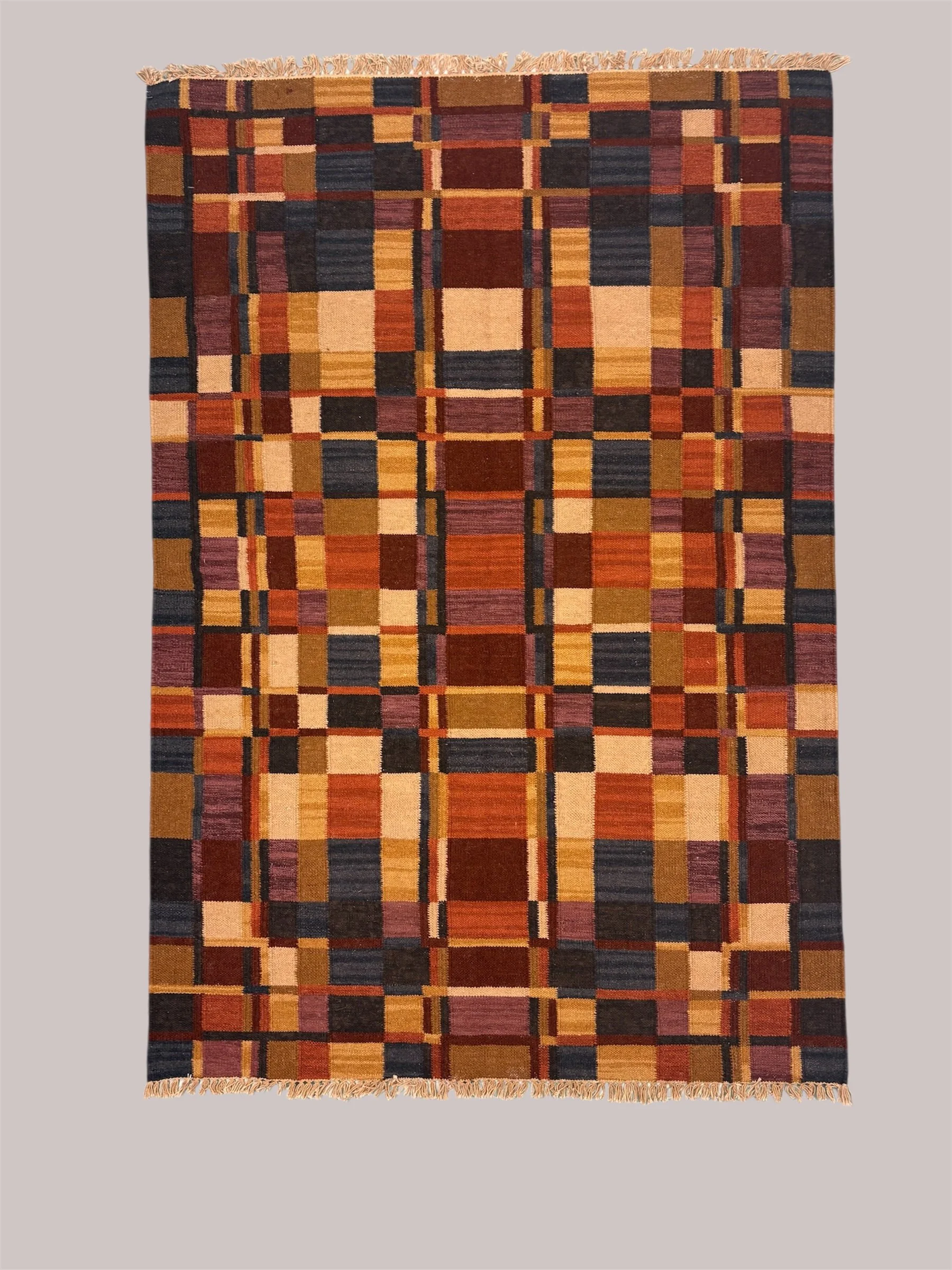 A rectangular area rug with a geometric pattern of squares and rectangles in warm tones of red, orange, brown, and yellow, with some black accents, and fringes at both ends.