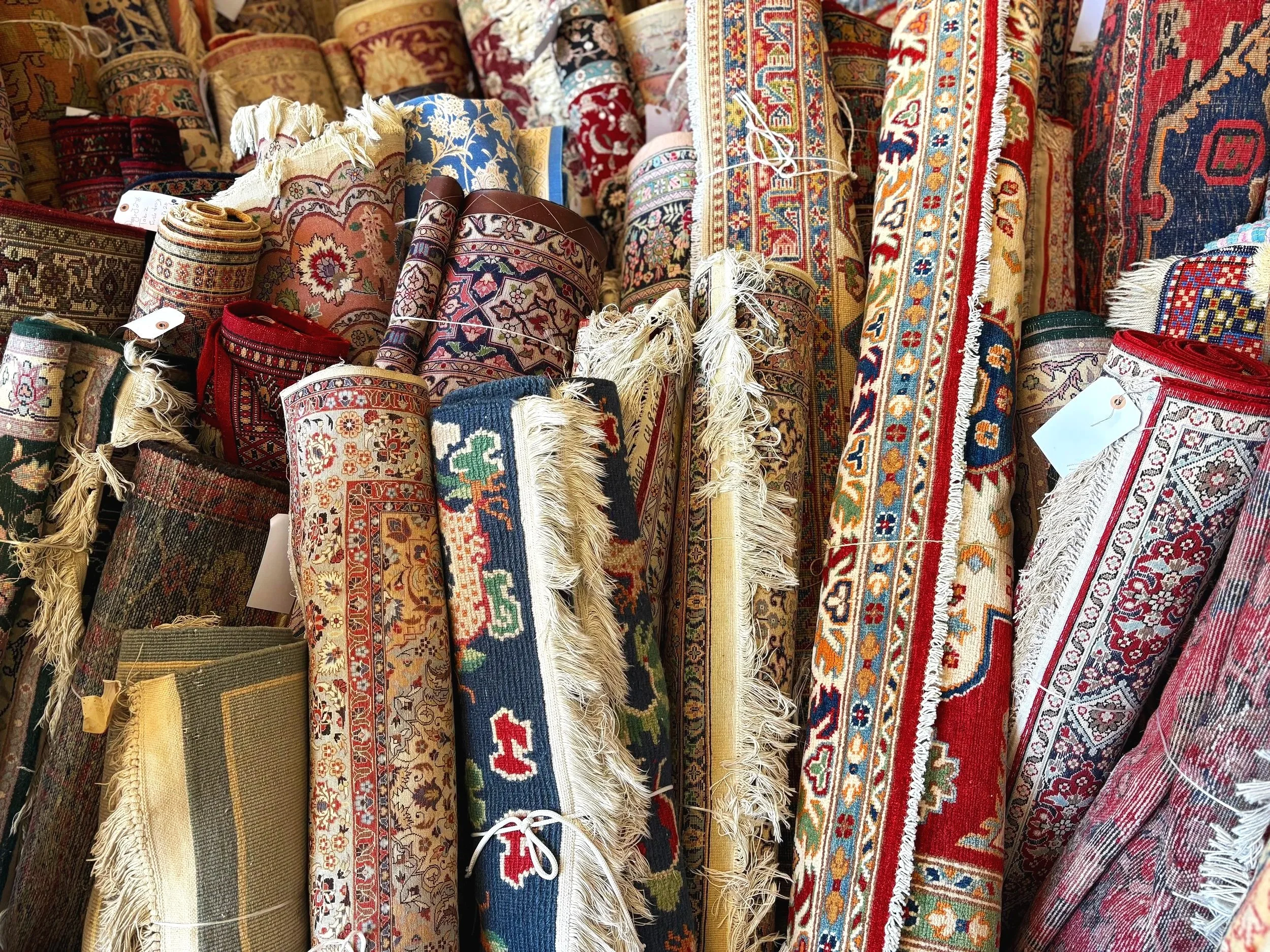 Stacked and rolled traditional patterned area rugs in various colors, including red, blue, beige, and green, displayed in a market or rug store.