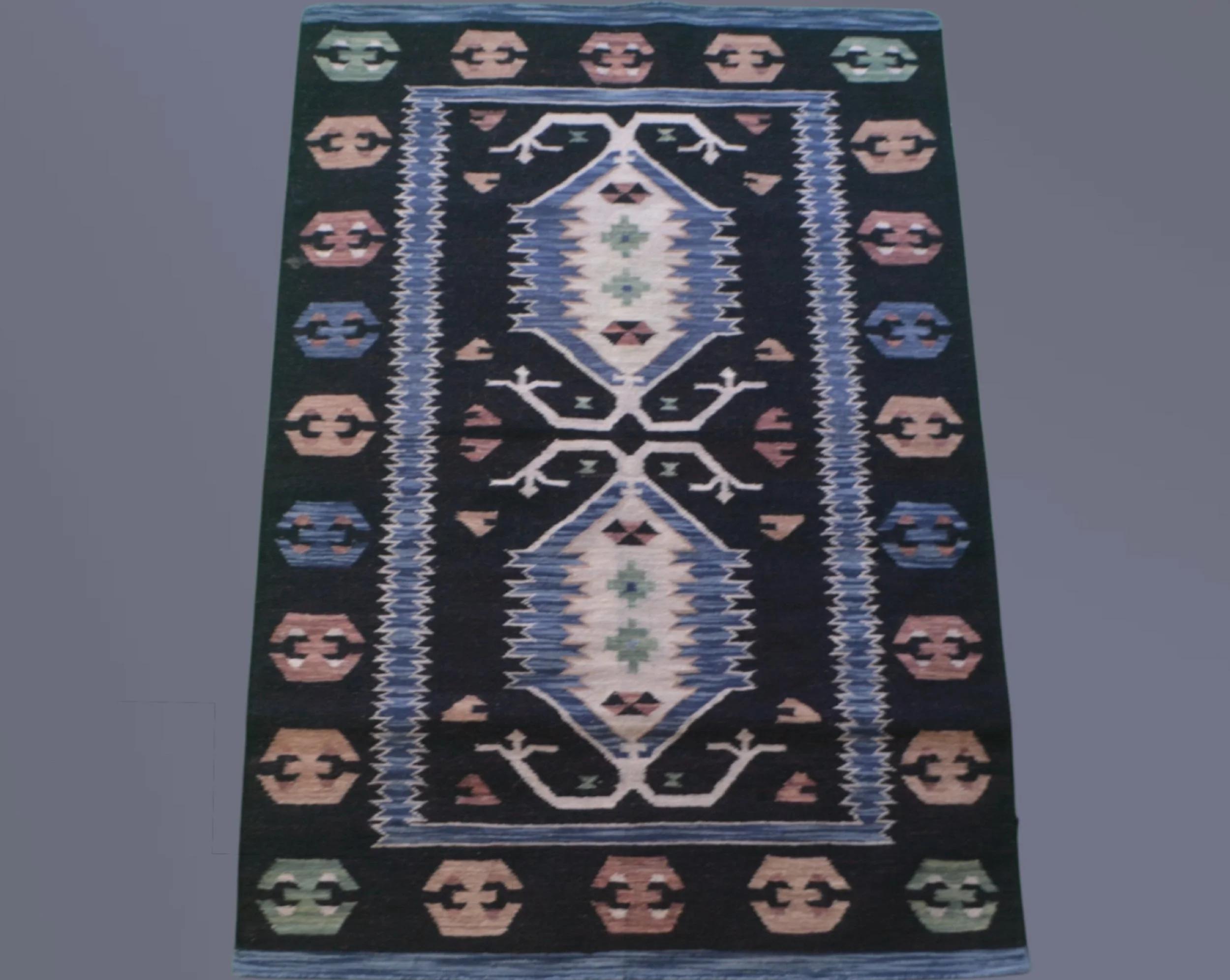 A rectangular area rug with a dark background, featuring a central diamond-shaped pattern in shades of blue, white, and green, bordered by stylized floral motifs in peach, blue, and green, with zigzag accents along the edges.