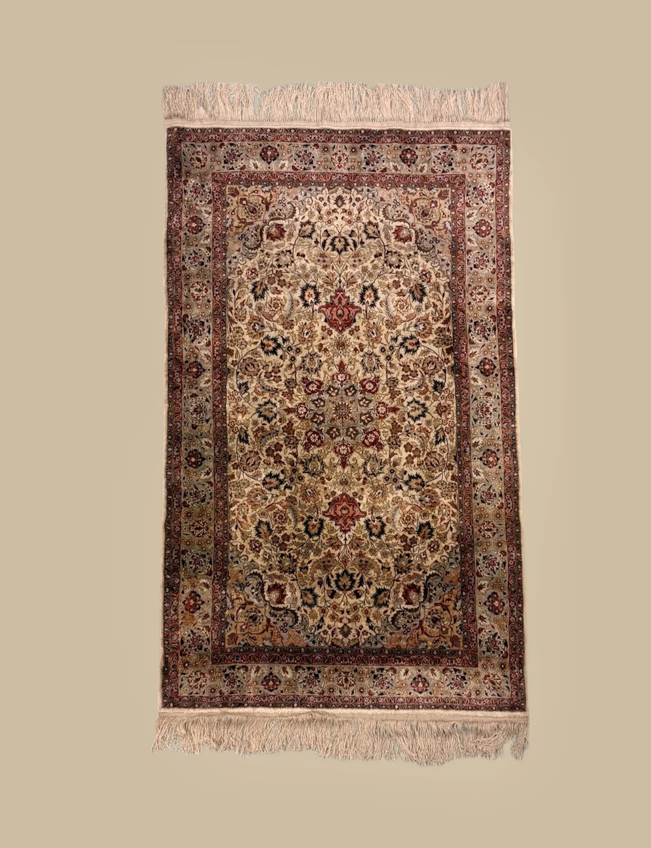 Traditional patterned area rug with floral and geometric designs in cream, red, and black colors, fringed edges, placed on a beige background.
